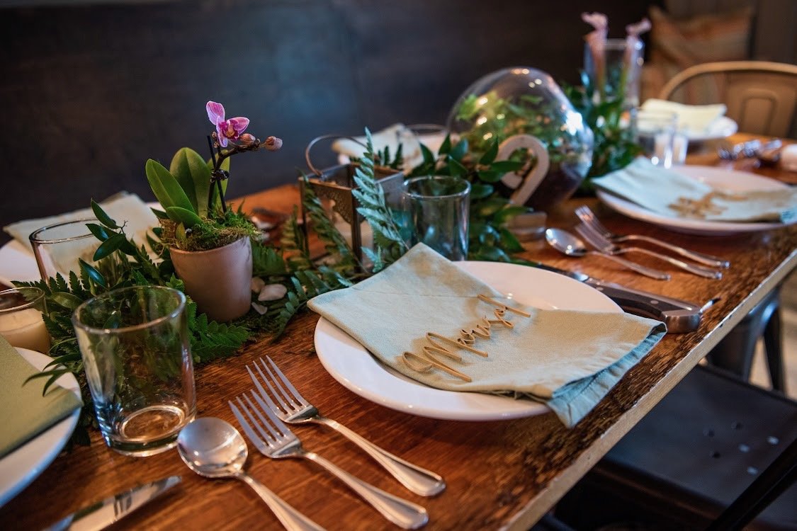 A decorated dining table with place settings, glassware, and a centerpiece of potted plants and greenery.