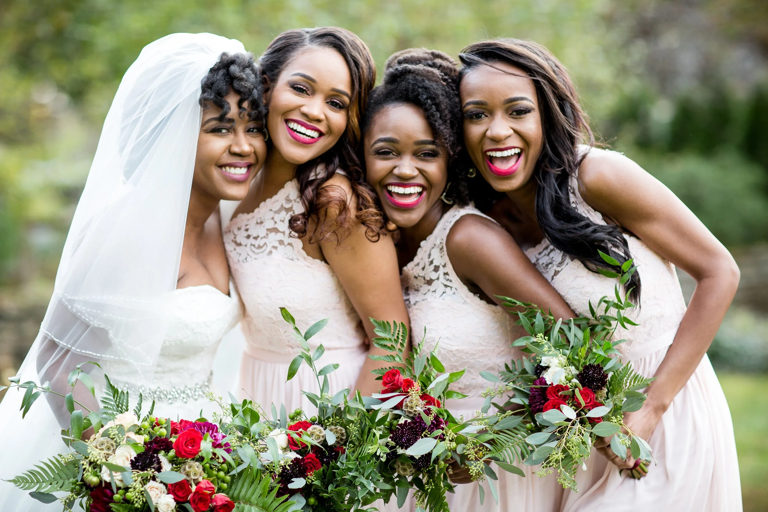 Four women smiling and embracing at an outdoor wedding, with a large floral bouquet in the foreground.