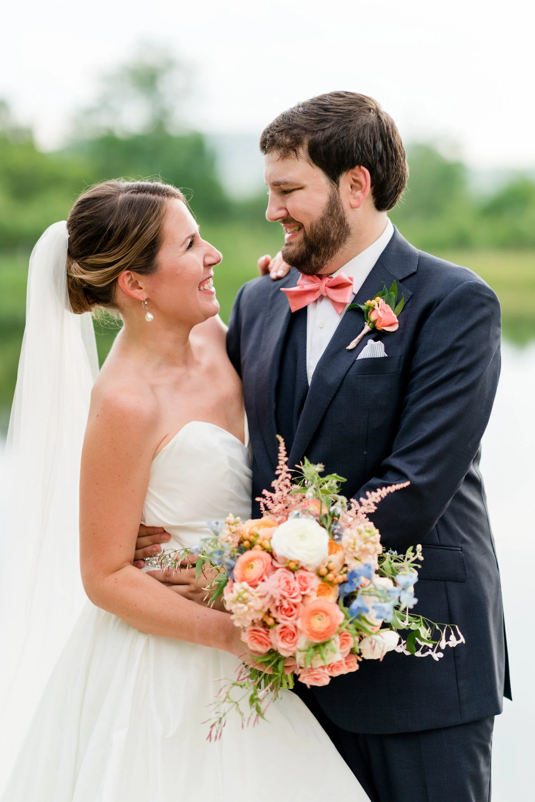 A bride and groom smiling and looking at each other outdoors, with the bride holding a colorful bouquet of flowers, in a wedding setting.