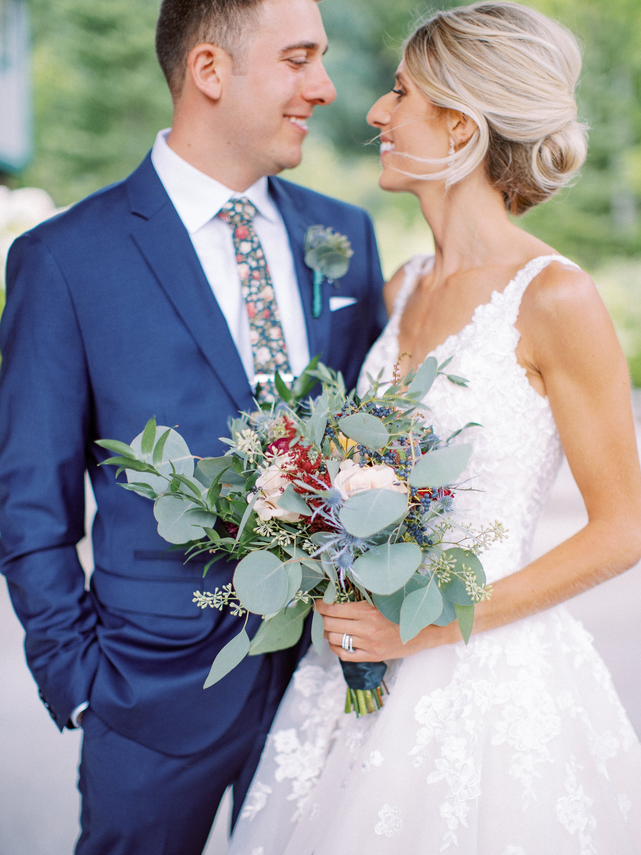 Close-up of a bride and groom smiling and facing each other, holding a bouquet of flowers with greenery, the bride wearing a lace wedding dress and the groom in a navy suit with a patterned tie.