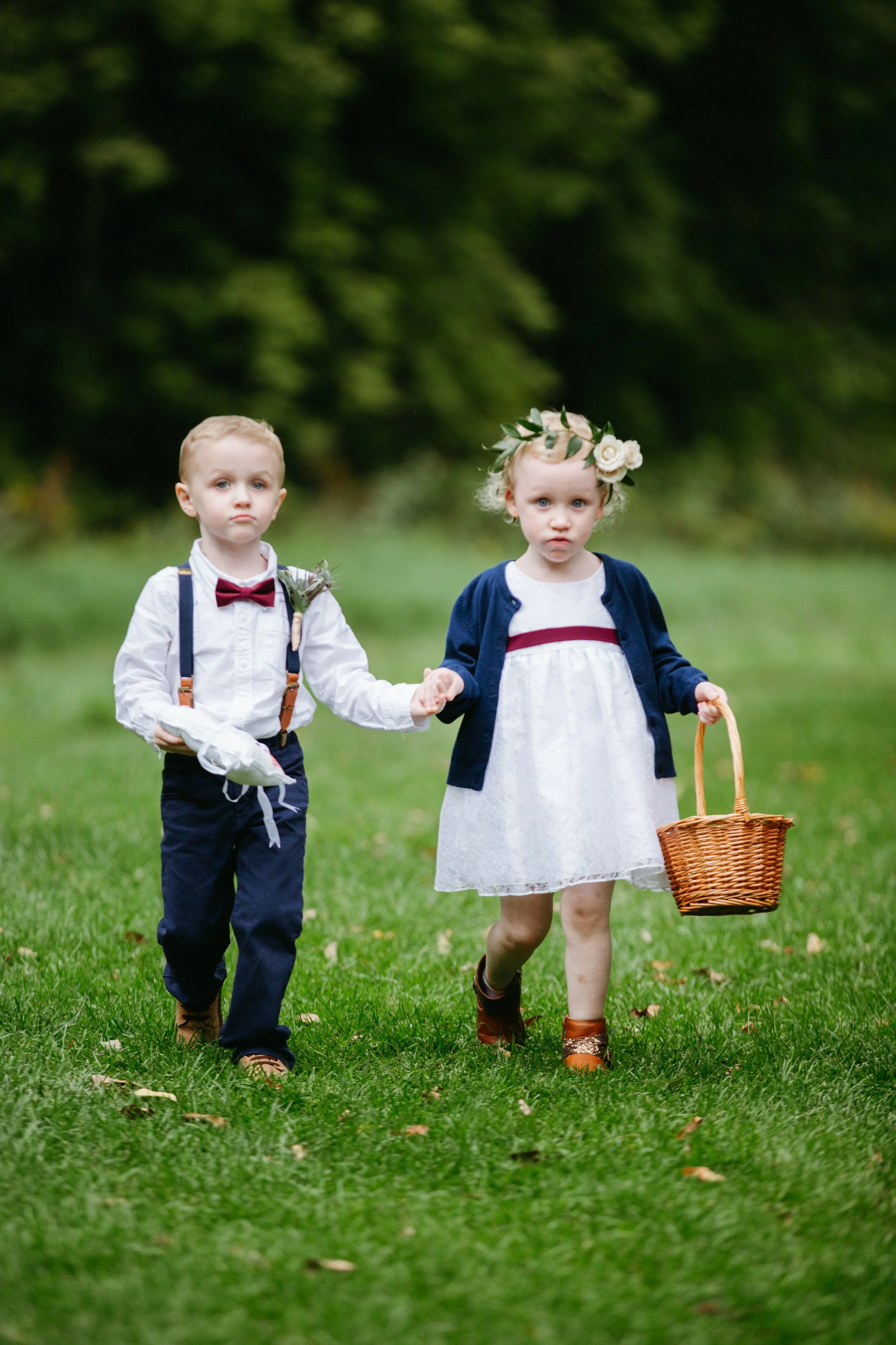 A young boy and girl holding hands, walking on a grassy field. The boy wears a white shirt, navy pants, suspenders, and a red bow tie. The girl wears a white dress with a navy cardigan, brown boots, and a flower crown. The girl holds a wicker basket.