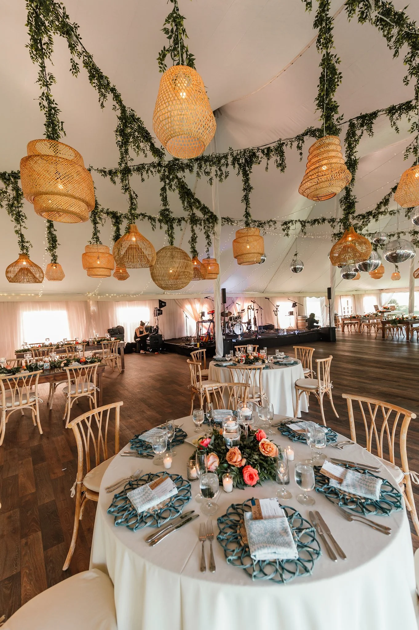 Elegant event reception with round tables covered in white tablecloths, decorated with floral centerpieces, candles, and place settings, set inside a large tent with hanging wicker pendant lights and green foliage.