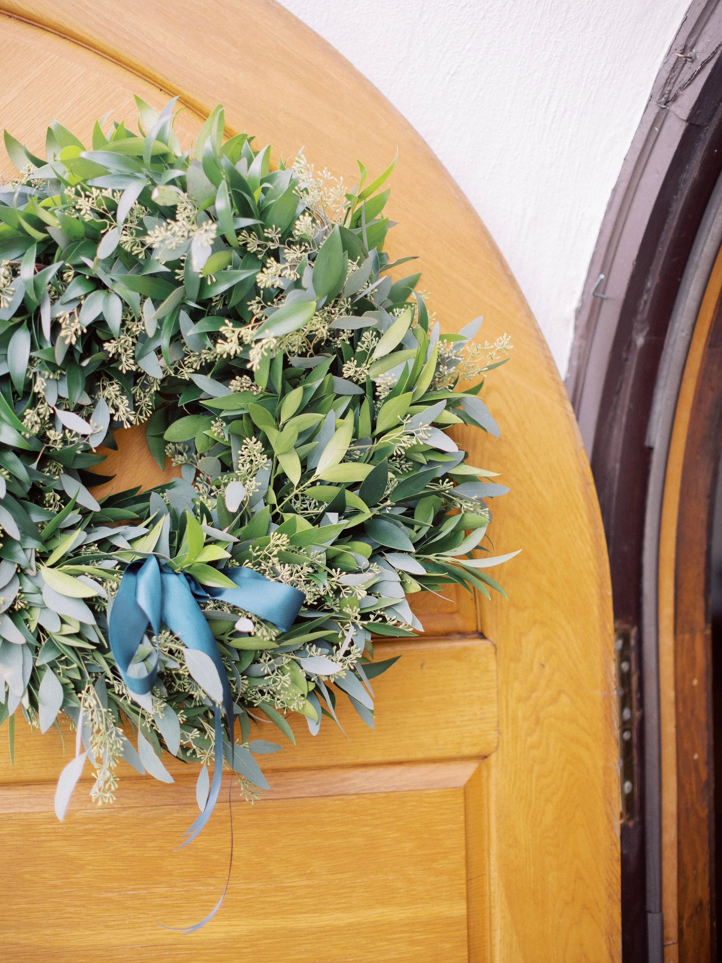 A wreath with green leaves and small white flowers hanging on a wooden door with a blue ribbon.