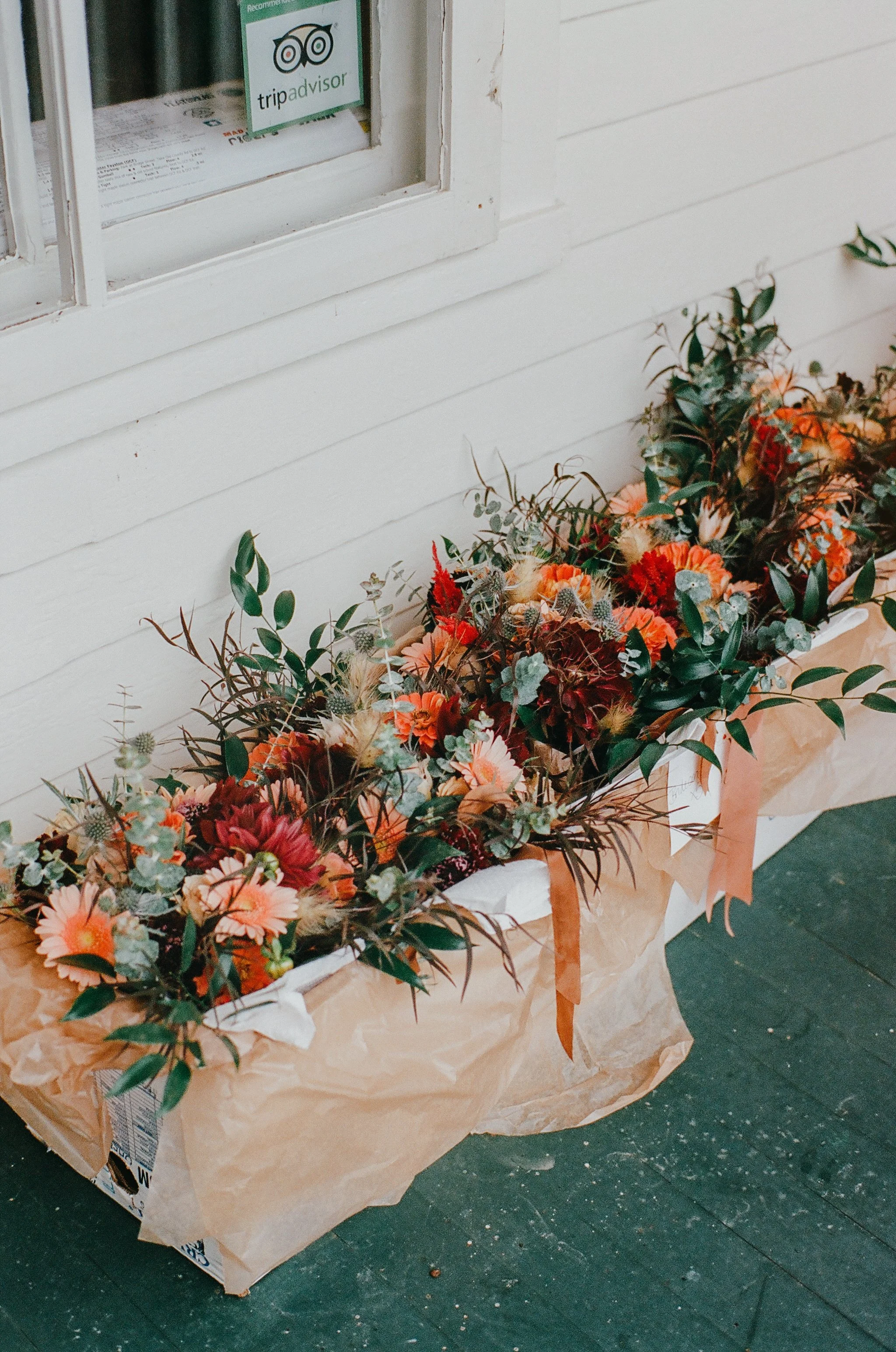 A large basket of colorful flowers, including orange, pink, and red blooms, with green foliage, wrapped in brown paper, placed on a black wooden surface near a white paneled wall and window with a TripAdvisor sticker.