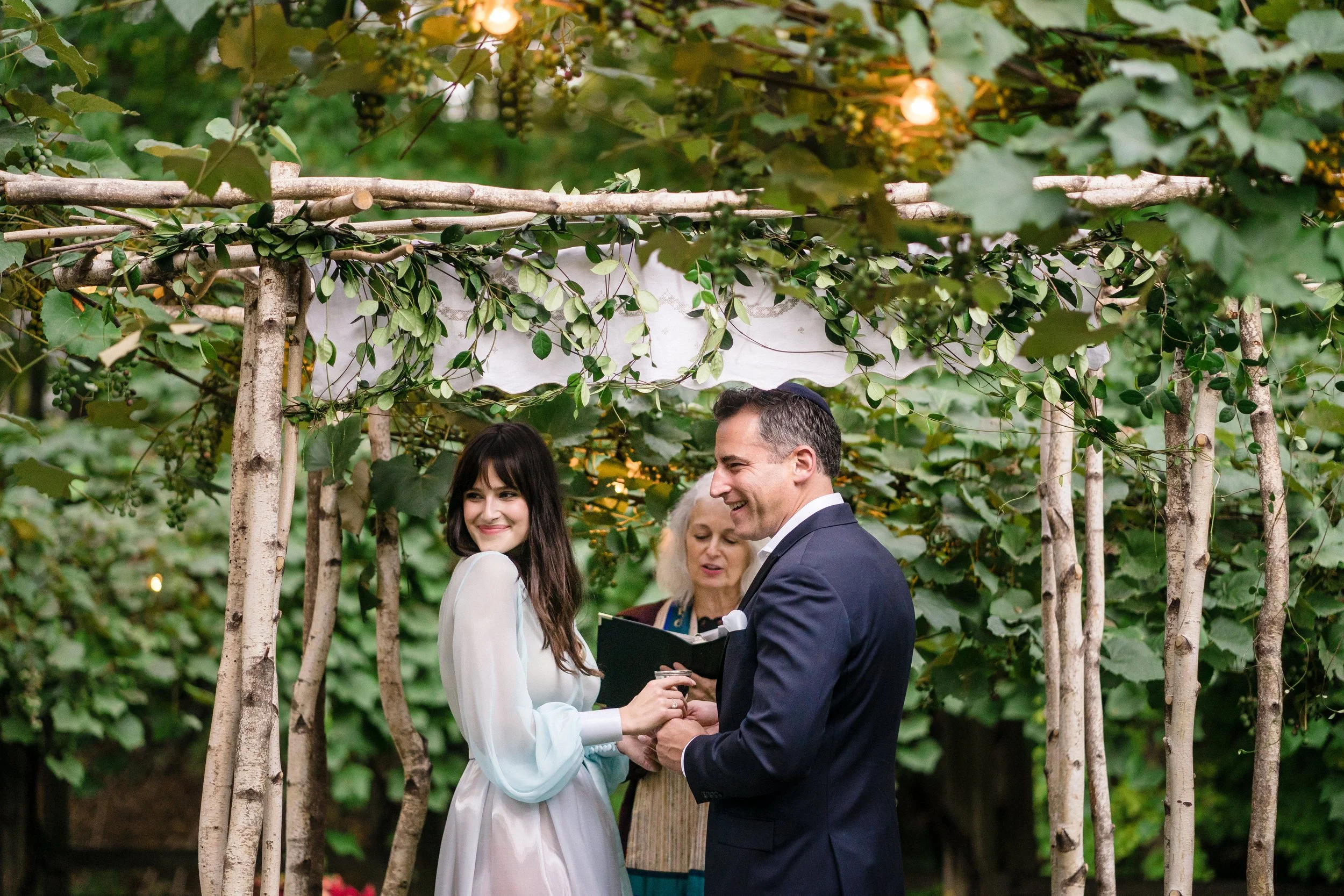 A bride and groom exchanging vows during their outdoor wedding ceremony, surrounded by greenery and decorated with wooden structures and string lights.