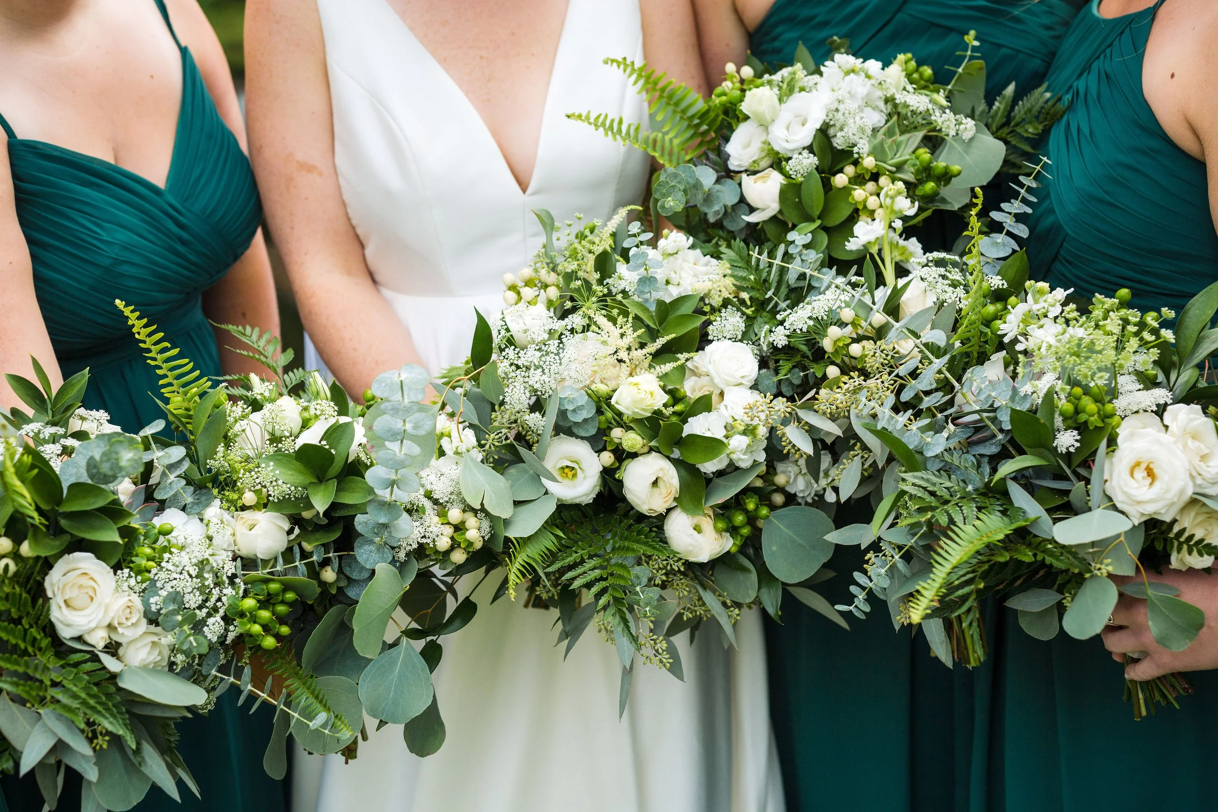 Close-up of a bride and bridesmaids holding a bouquet of white flowers and green foliage at a wedding.