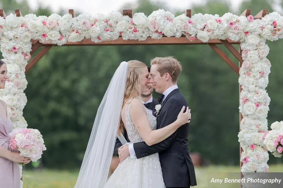 A bride and groom share a kiss during their wedding ceremony under a floral archway in an outdoor setting.