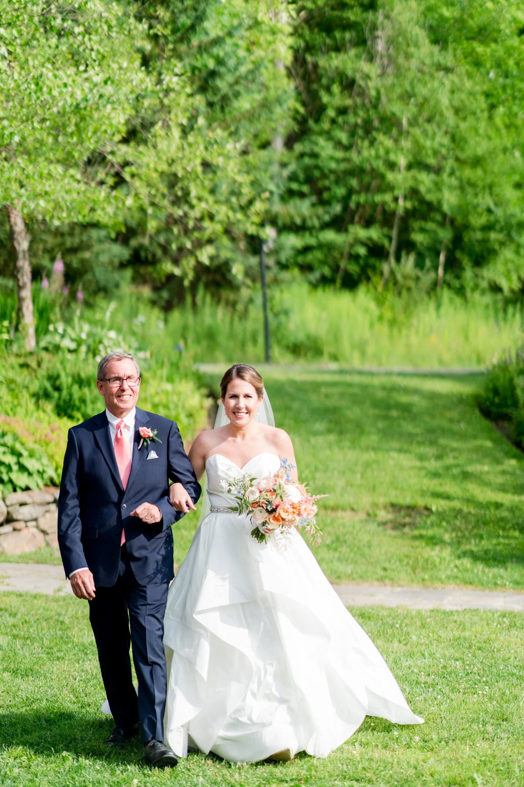 A bride in a white wedding dress holding a bouquet of flowers walking beside a man in a navy suit with glasses outdoors in a park with green trees.