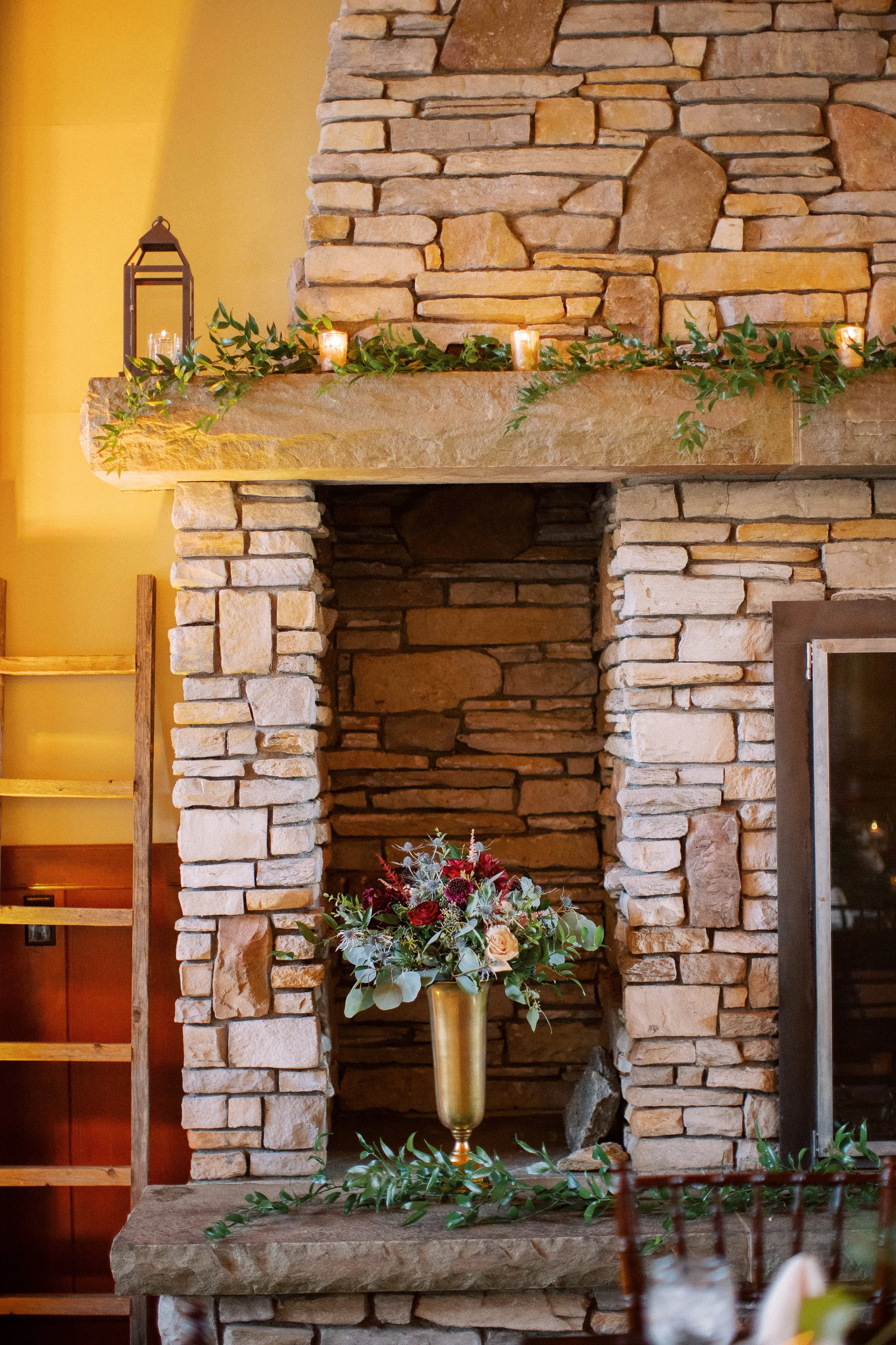 A stone fireplace with a wood mantle decorated with candles, greenery, and a lantern. There is a floral arrangement in a gold vase on the fireplace hearth.