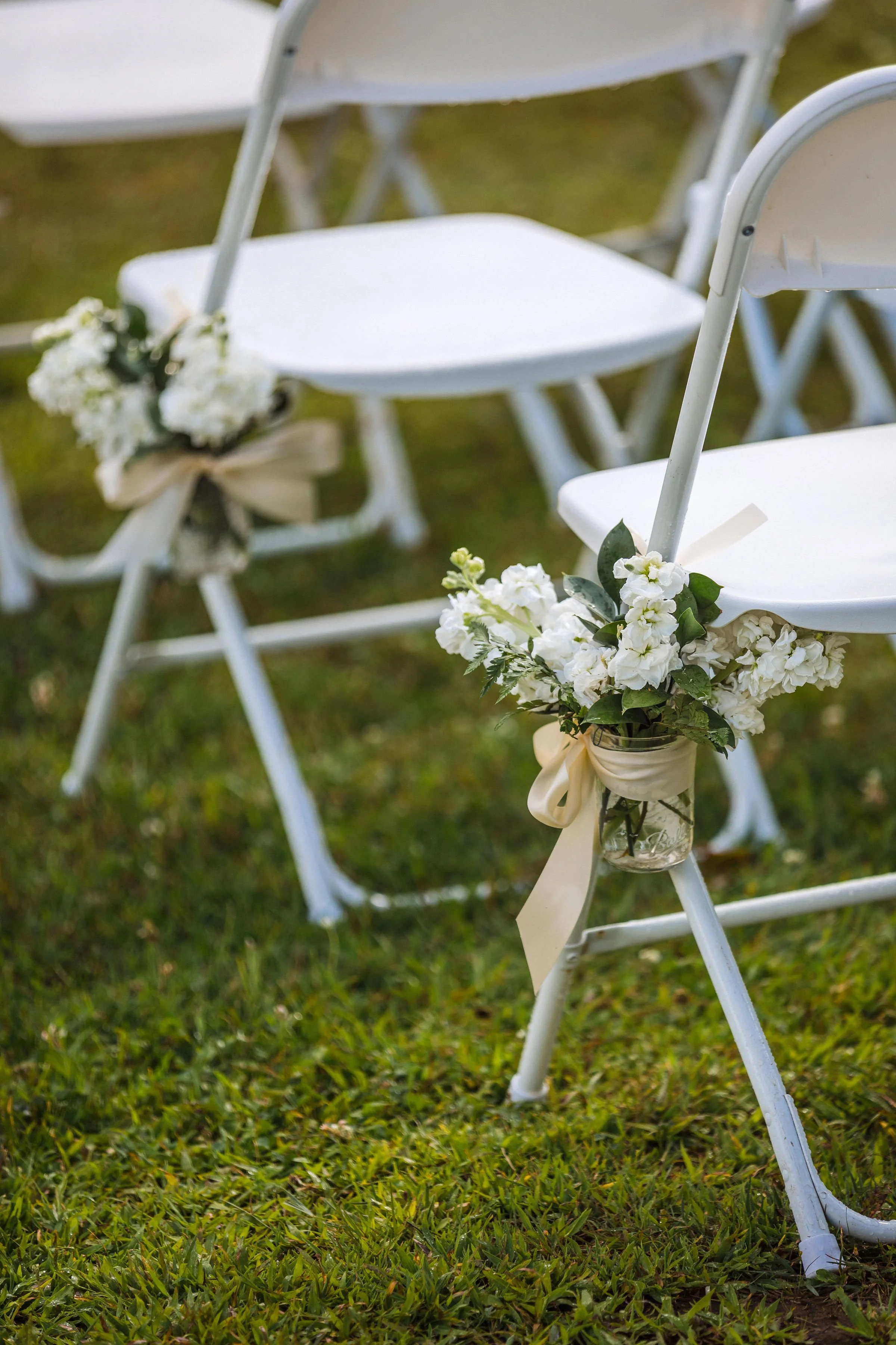 White chairs decorated with bouquets of white flowers and beige ribbons on a grassy outdoor setting, likely for a wedding or special event.