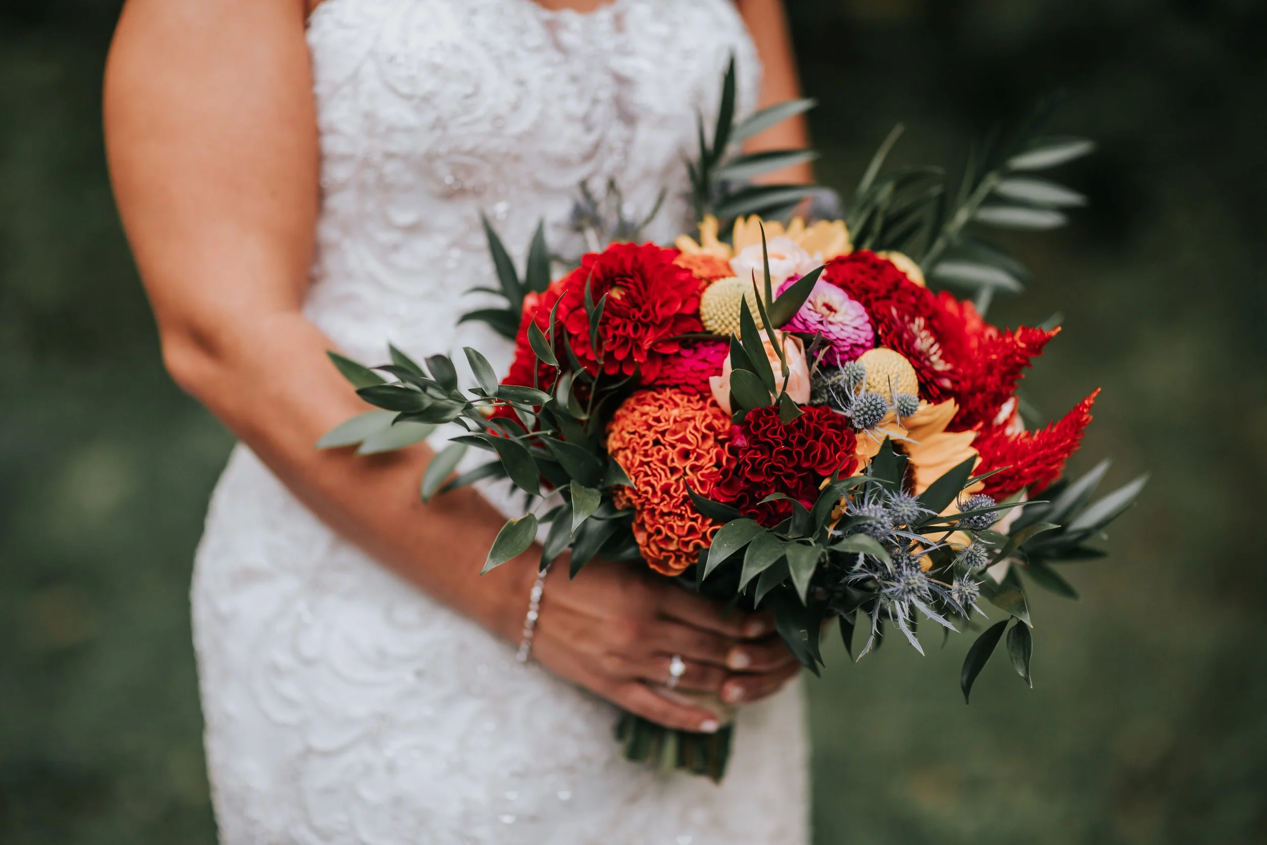 A woman in a lace white dress holding a colorful bouquet of flowers, including red, orange, yellow, and purple blooms, and green leaves, outdoors.