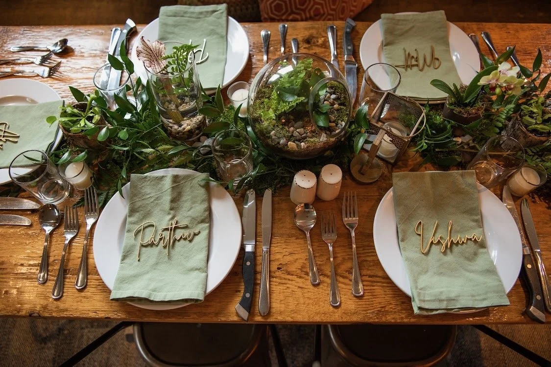 A rustic wooden dining table set for a meal with four place settings, green cloth napkins, and name tags reading 'Patience', 'Yeshua', and others. The table has green foliage, a glass terrarium with plants, and small candles as decorations.