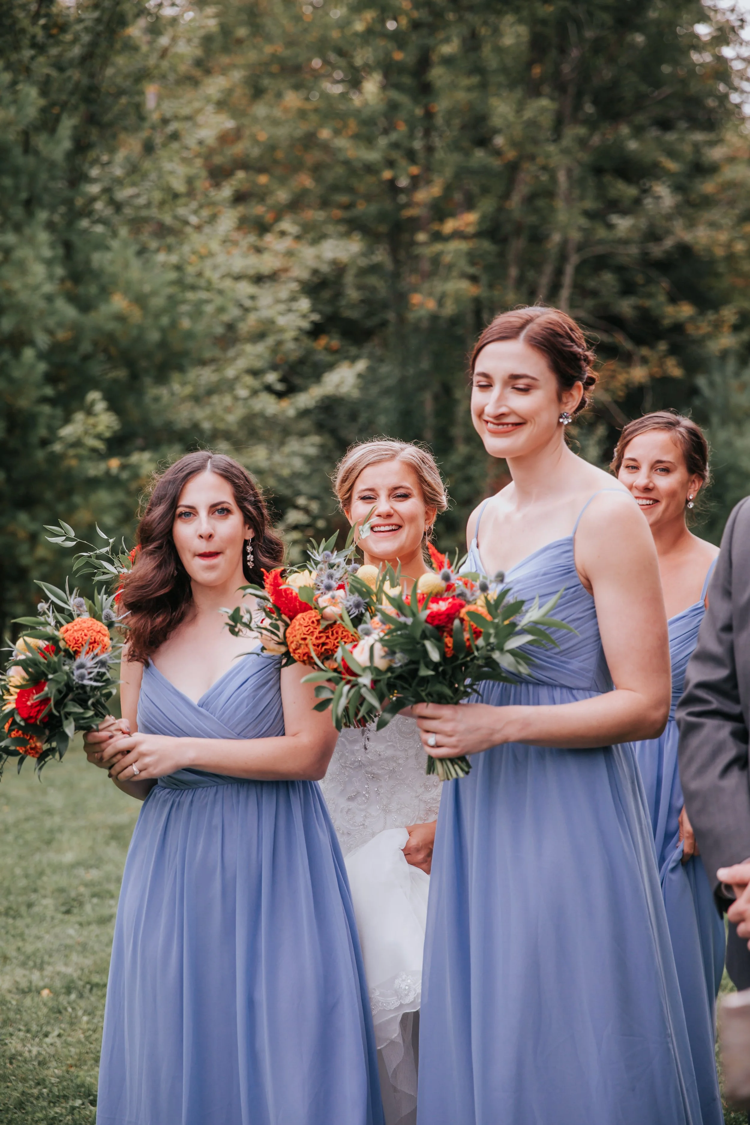 A group of women in blue bridesmaid dresses holding bouquets of flowers, standing outdoors with trees in the background.