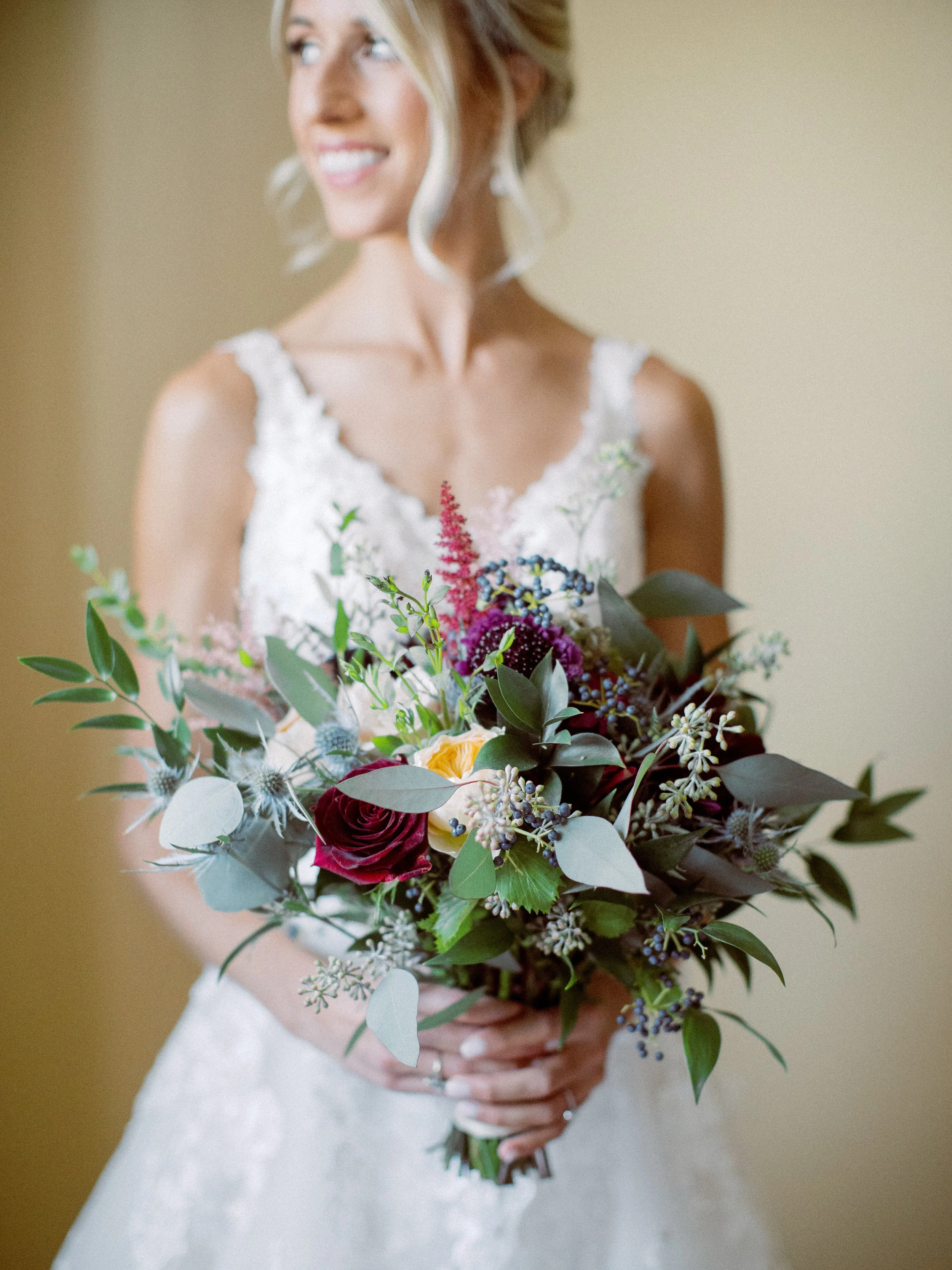 A woman in a white wedding dress holding a large bouquet of colorful flowers, smiling and looking to her right.