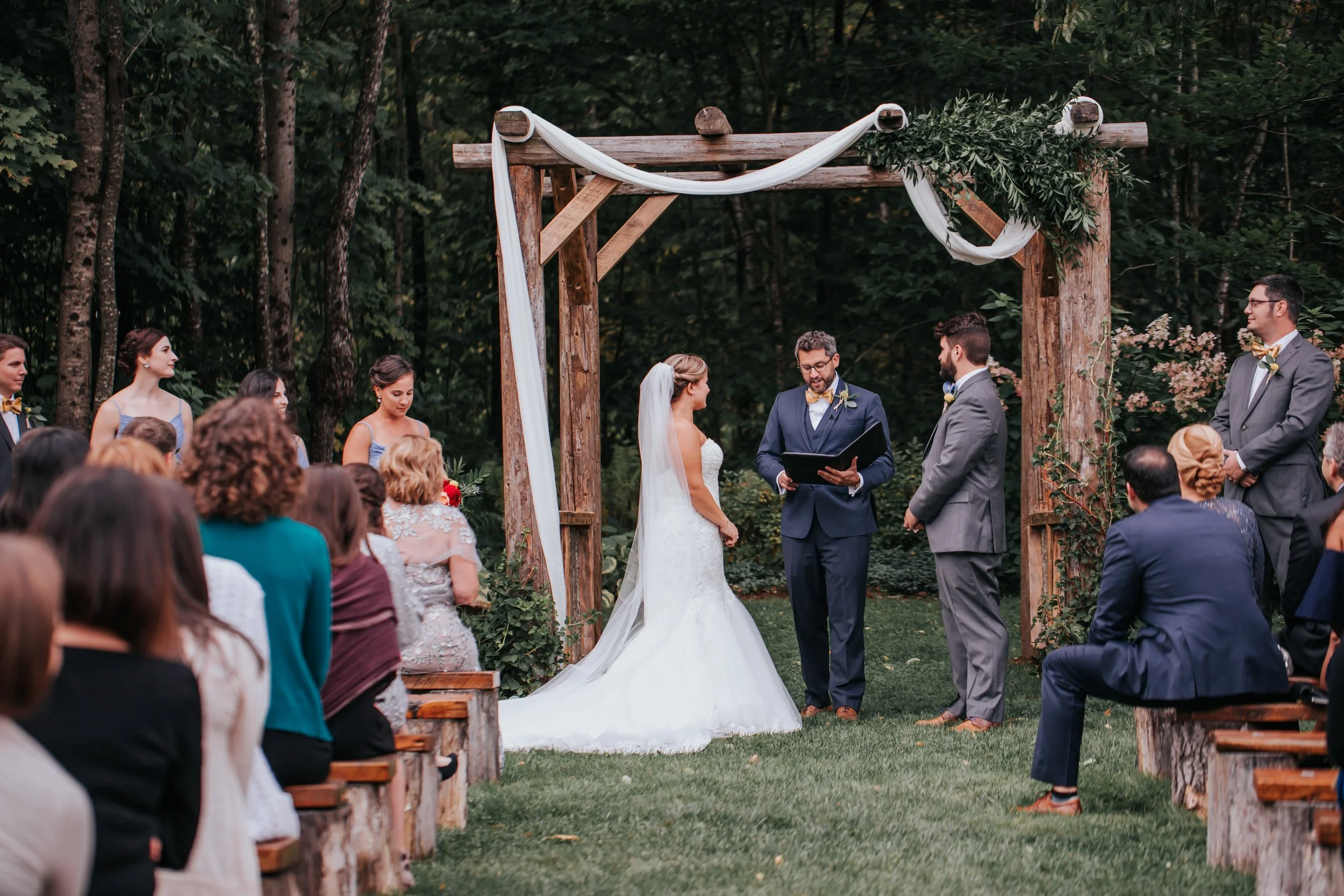 A couple getting married outdoors under a wooden arch decorated with white fabric and greenery, surrounded by guests in a forest setting.