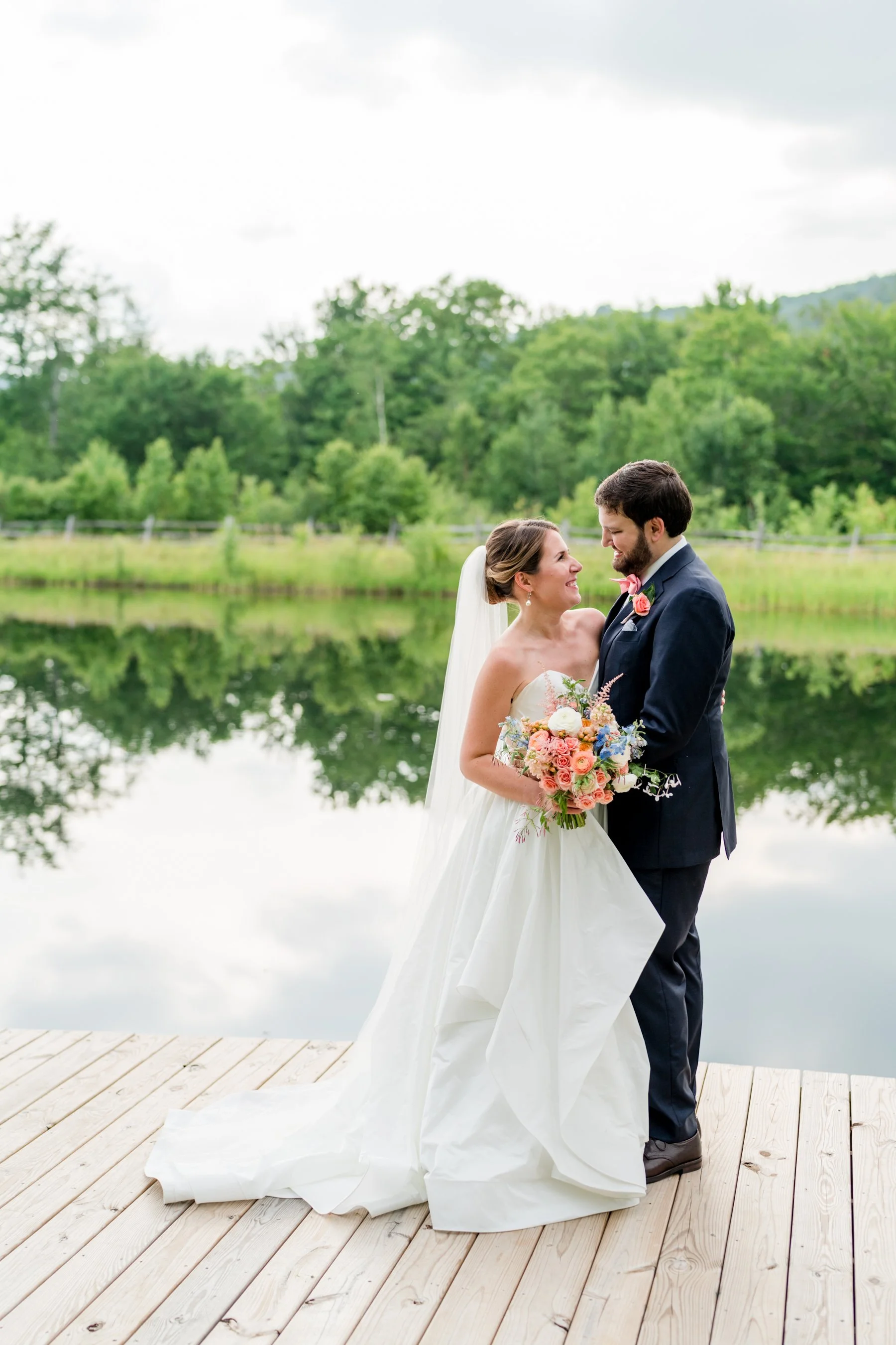 A bride and groom standing on a wooden dock by a lake, smiling at each other. The bride is holding a bouquet of pink, peach, and white flowers. The background features green trees and a cloudy sky.