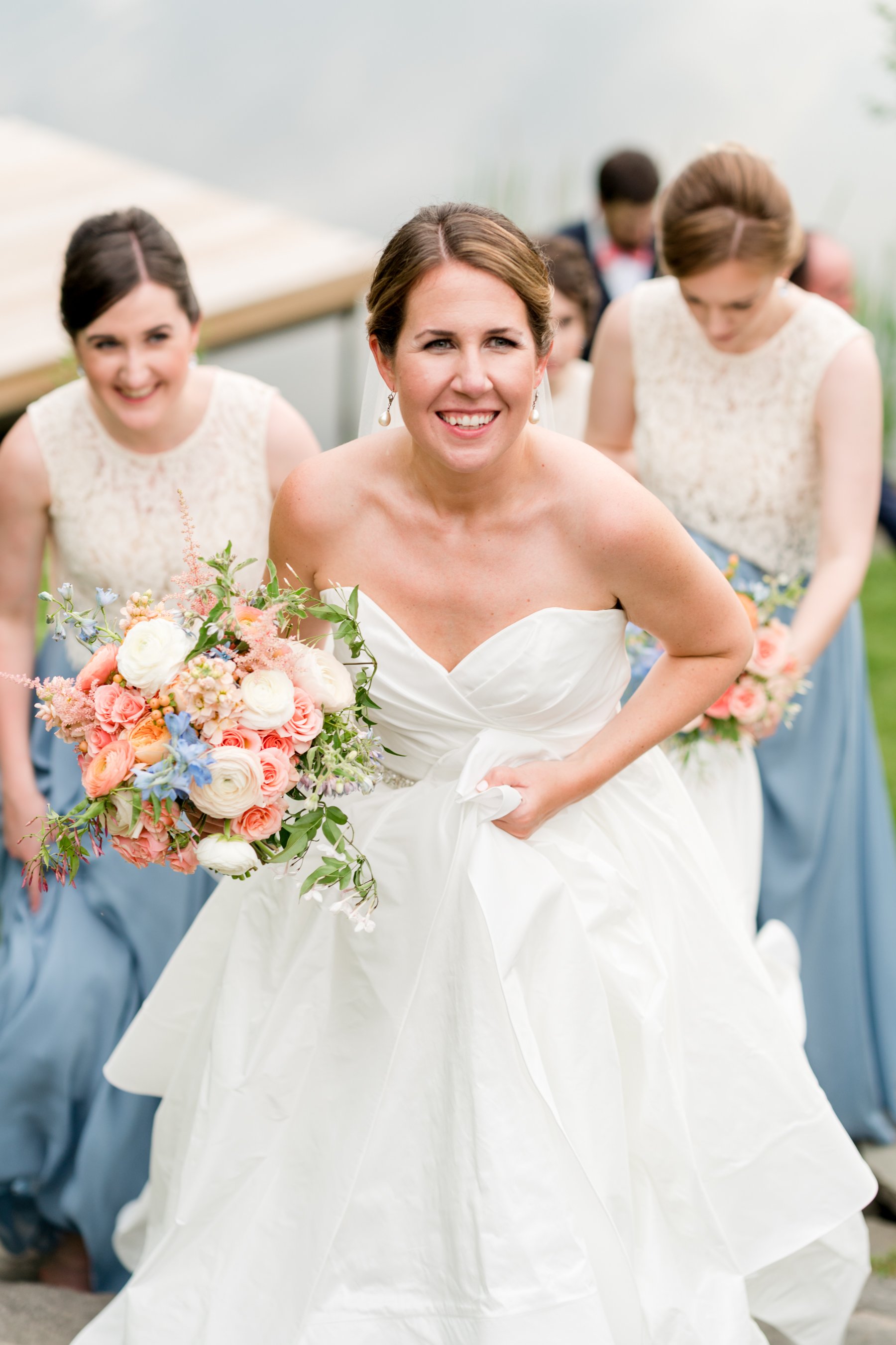 A bride in a white wedding dress holding a bouquet of mixed flowers, smiling, surrounded by bridesmaids in light blue dresses during a wedding celebration.