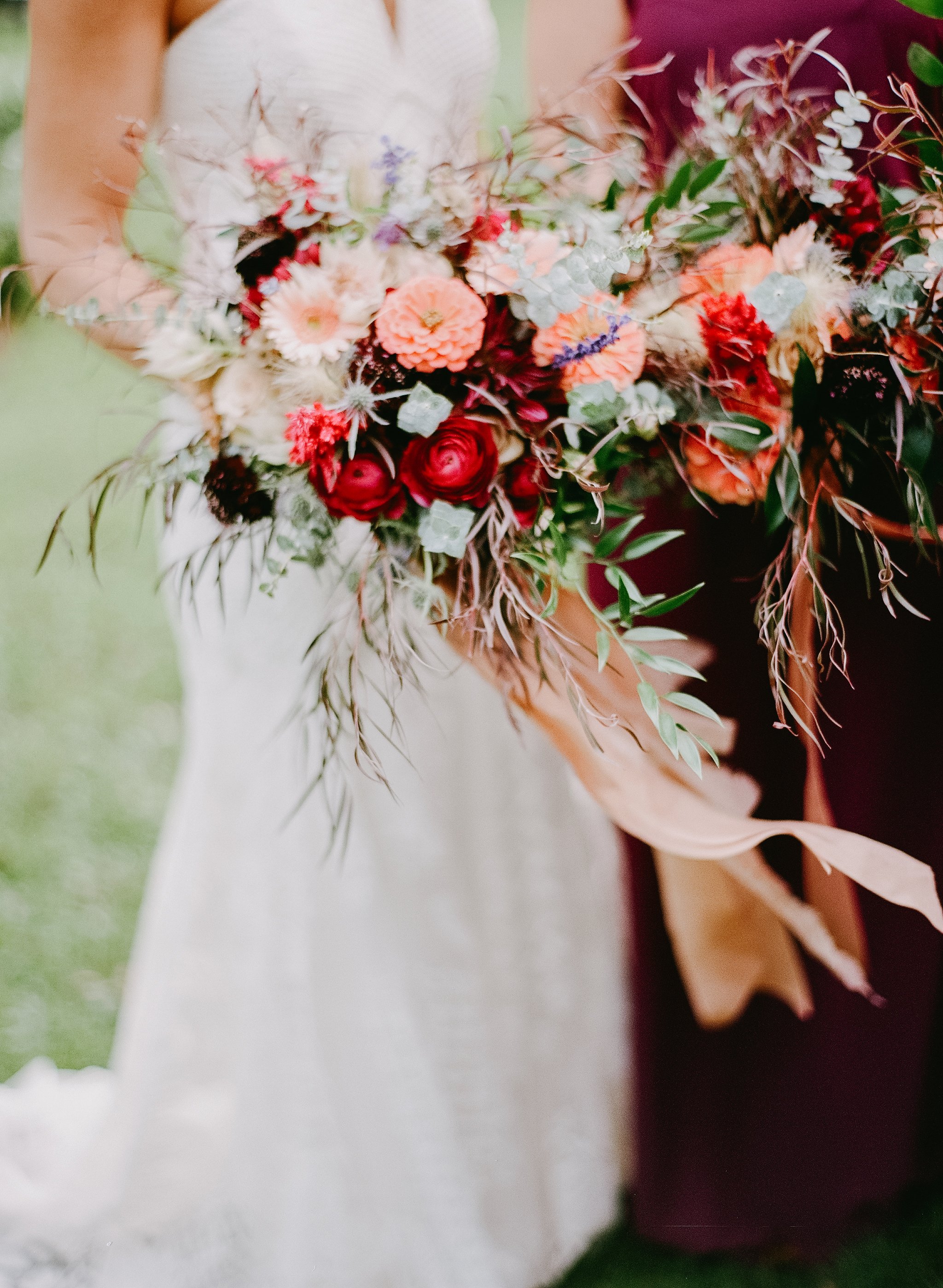 Close-up of a bouquet of mixed flowers, including red roses, peach-colored dahlias, white and purple small flowers, greenery, and dried foliage, held by a person wearing a light-colored dress.
