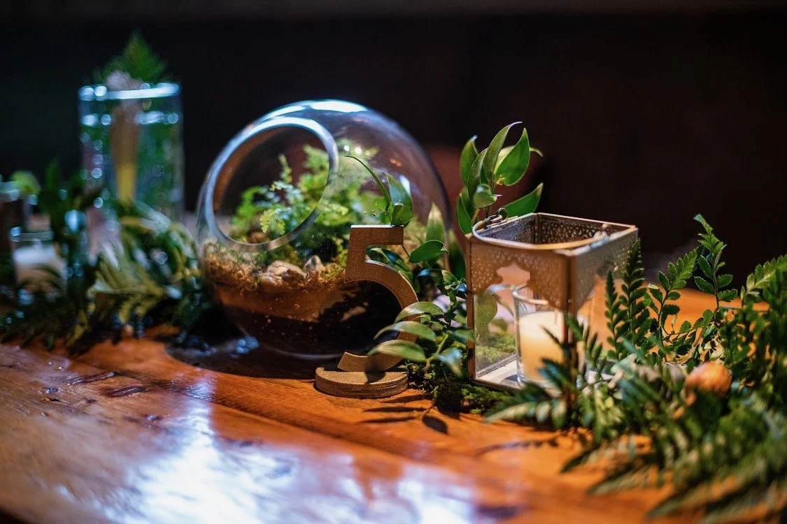 Decorative indoor scene with glass terrarium, green plants, a candle in a metallic holder, a wooden number 5, and greenery on a wooden surface.