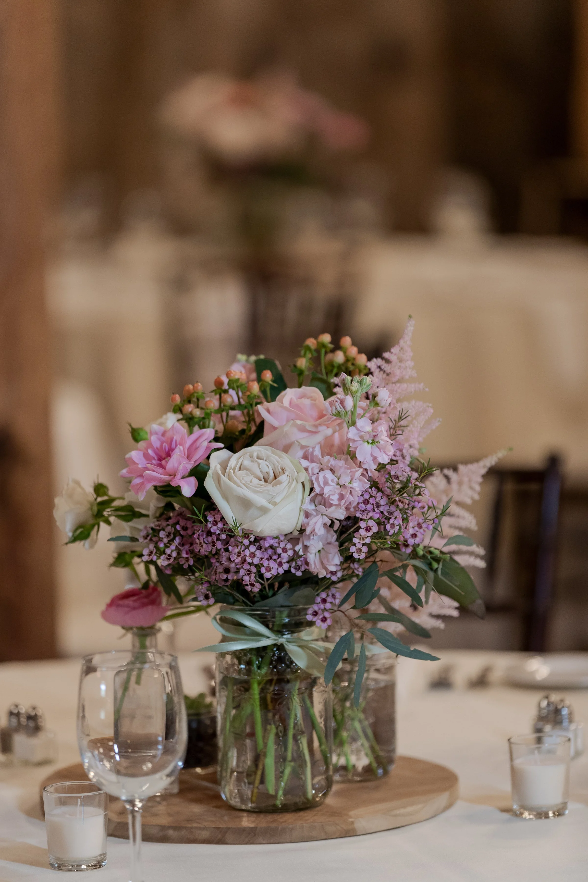 A floral arrangement with pink, white, and purple flowers in a glass vase on a round wooden table surrounded by wine glasses and candles.