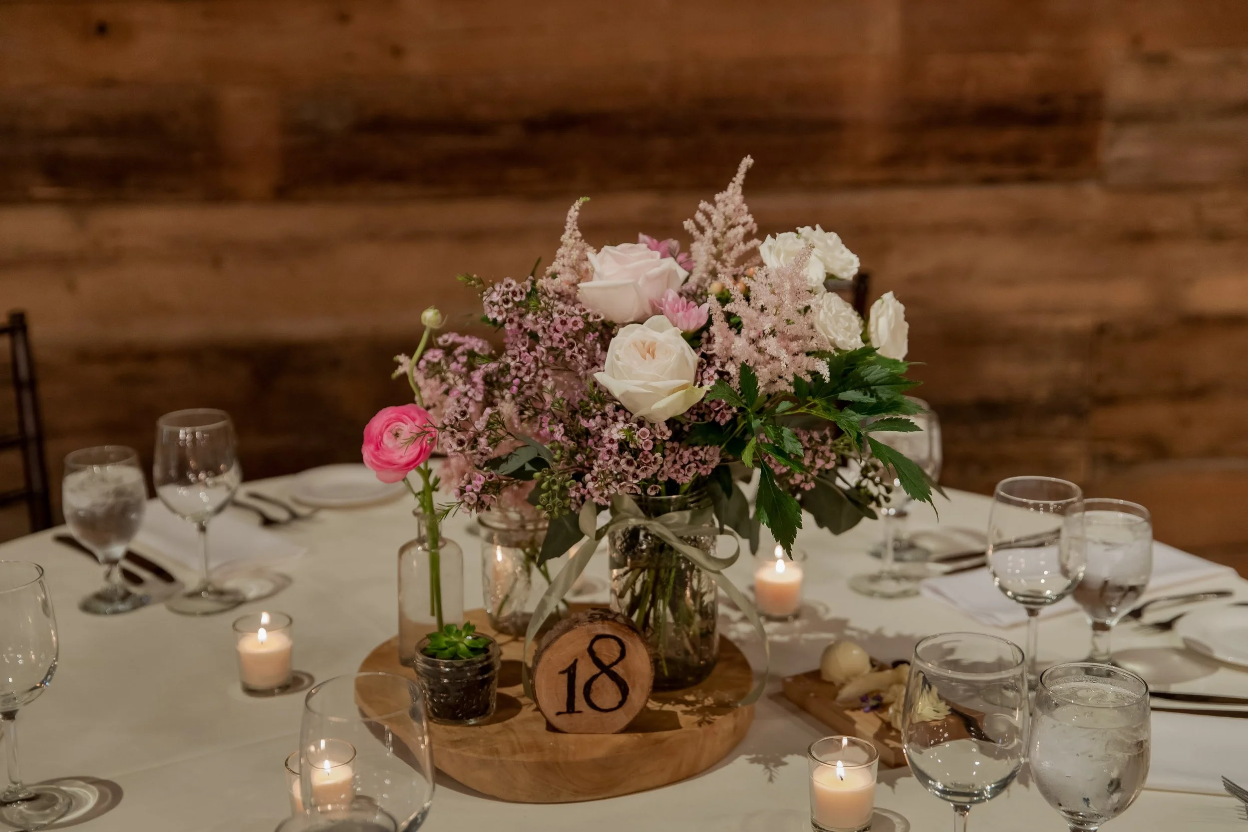 A floral centerpiece with pink and white flowers in a glass jar, surrounded by lit candles on a decorated table for a celebration, with a wooden wall in the background.