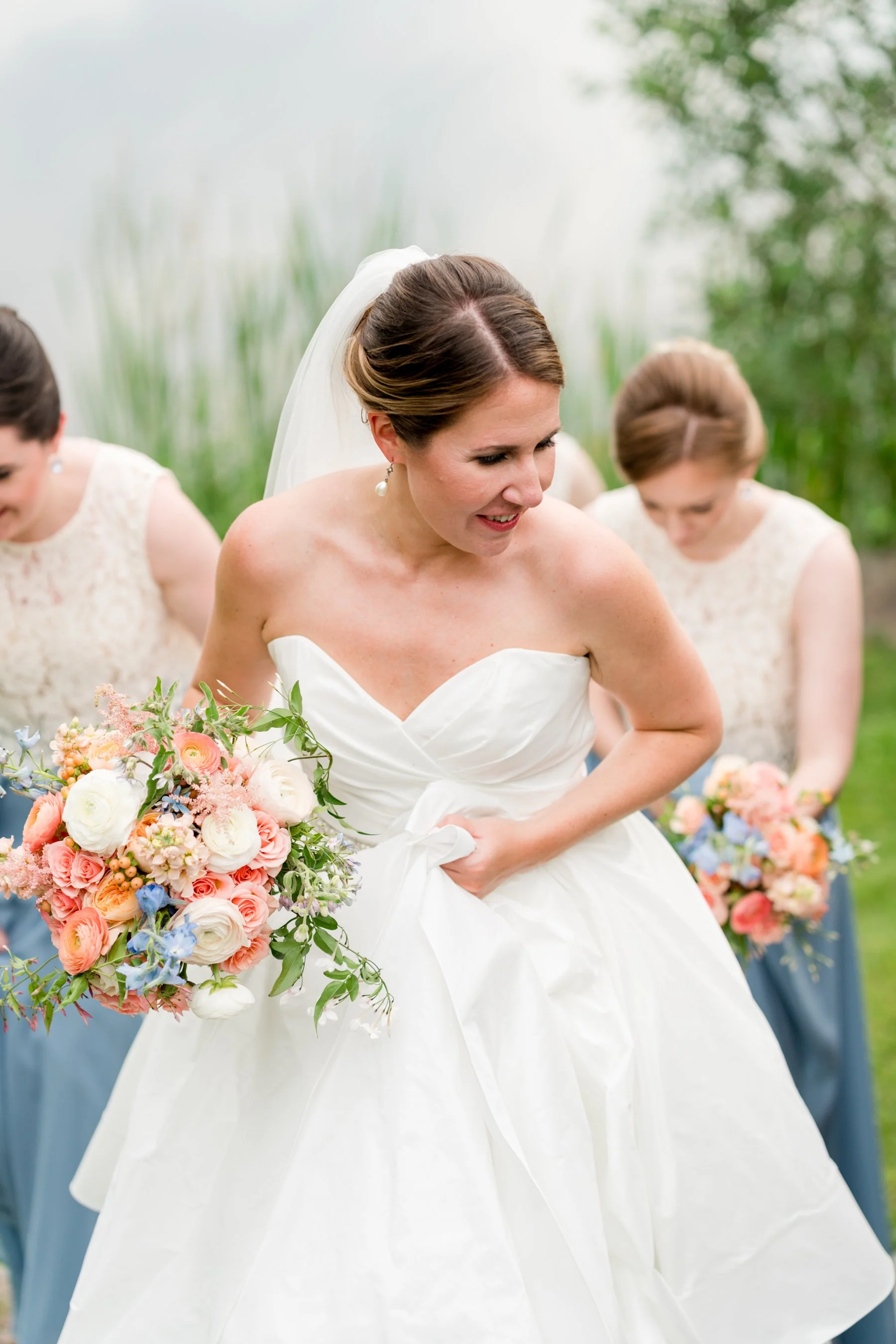 Bride in a white wedding dress holding a bouquet of pink, white, and blue flowers, surrounded by bridesmaids in light-colored dresses outdoors.