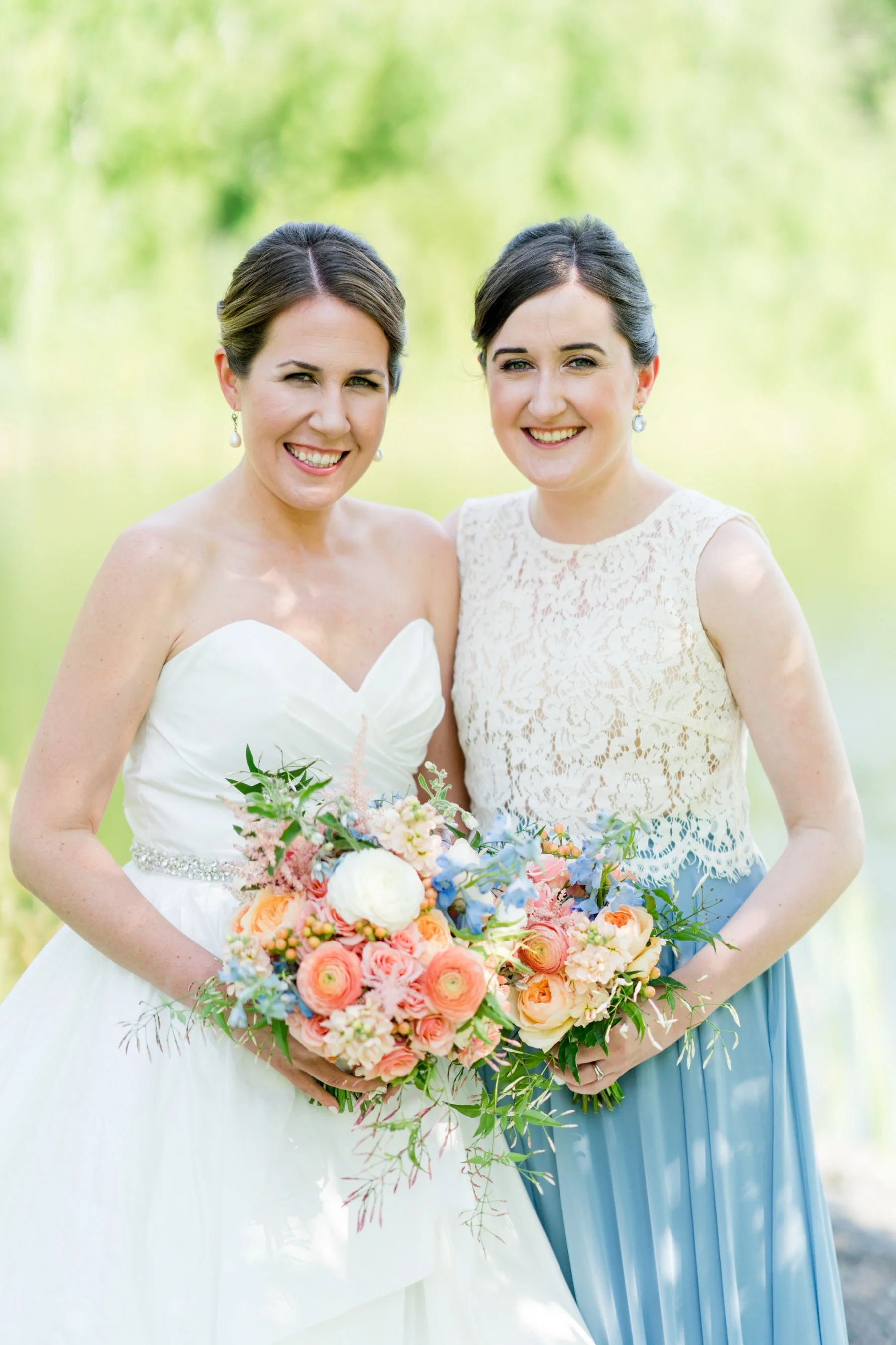 Two women, one dressed in a wedding gown and the other in a pastel blue dress, stand outdoors with a blurred green background. The woman in the wedding gown holds a large bouquet of colorful flowers.