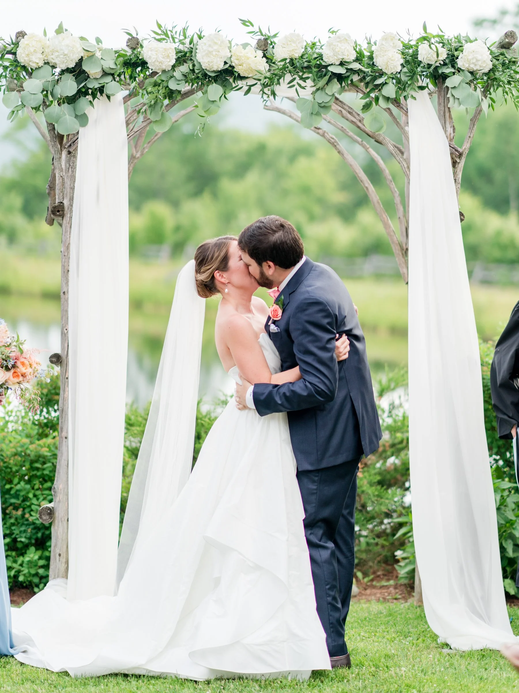 A couple in wedding attire sharing a kiss under a floral wedding arch outdoors with green trees and grass in the background.