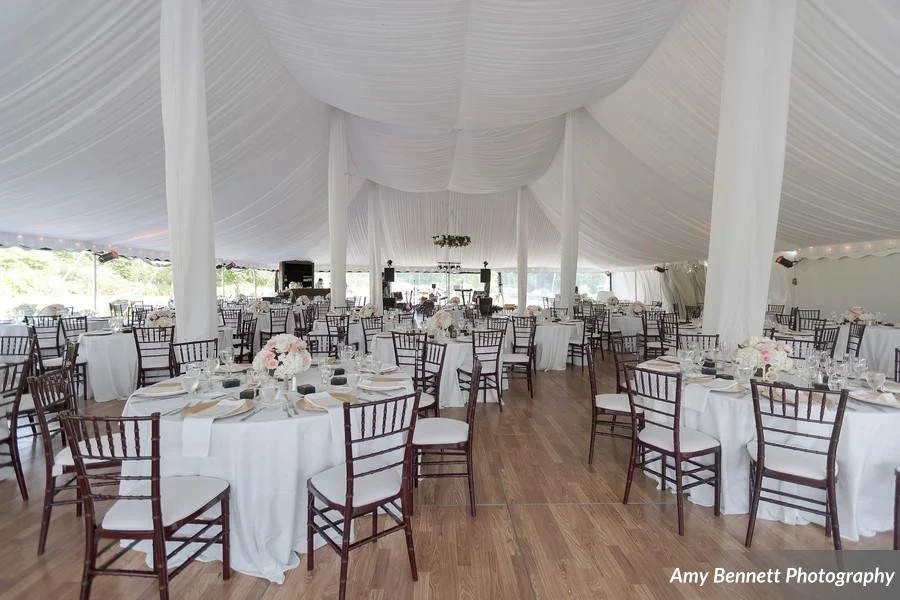 Interior of a spacious event tent set up for a wedding or celebration, with round tables decorated with white tablecloths, floral centerpieces, and dark wood chairs, with a wooden floor and natural light coming in.