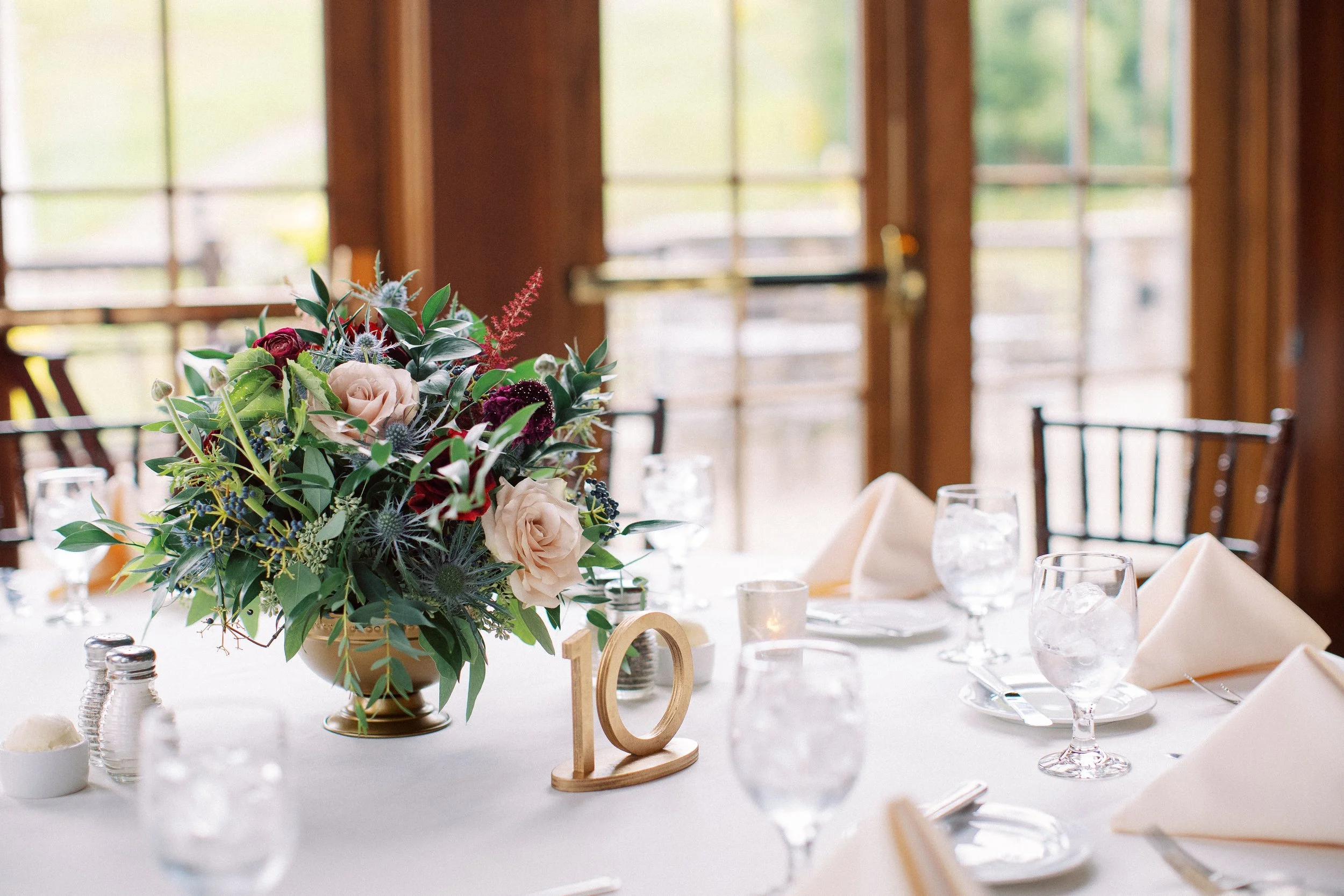 A table decorated with a floral centerpiece of pink, purple, and white flowers with greenery, set for a formal event in a bright room with large windows and wooden frames.
