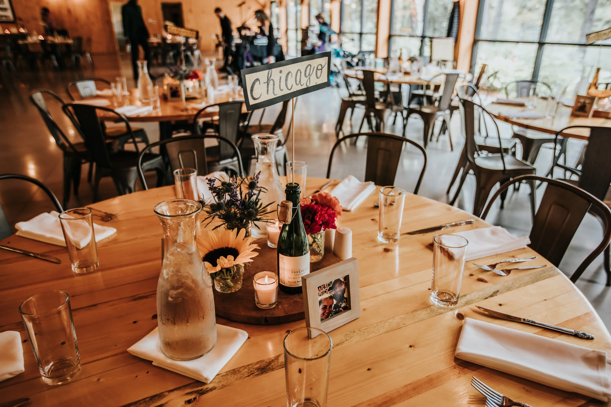 A decorated dining table set for an event with a 'Chicago' place card, floral centerpiece, wine bottle, candles, glasses, and napkins in a restaurant with large windows and wooden interior.