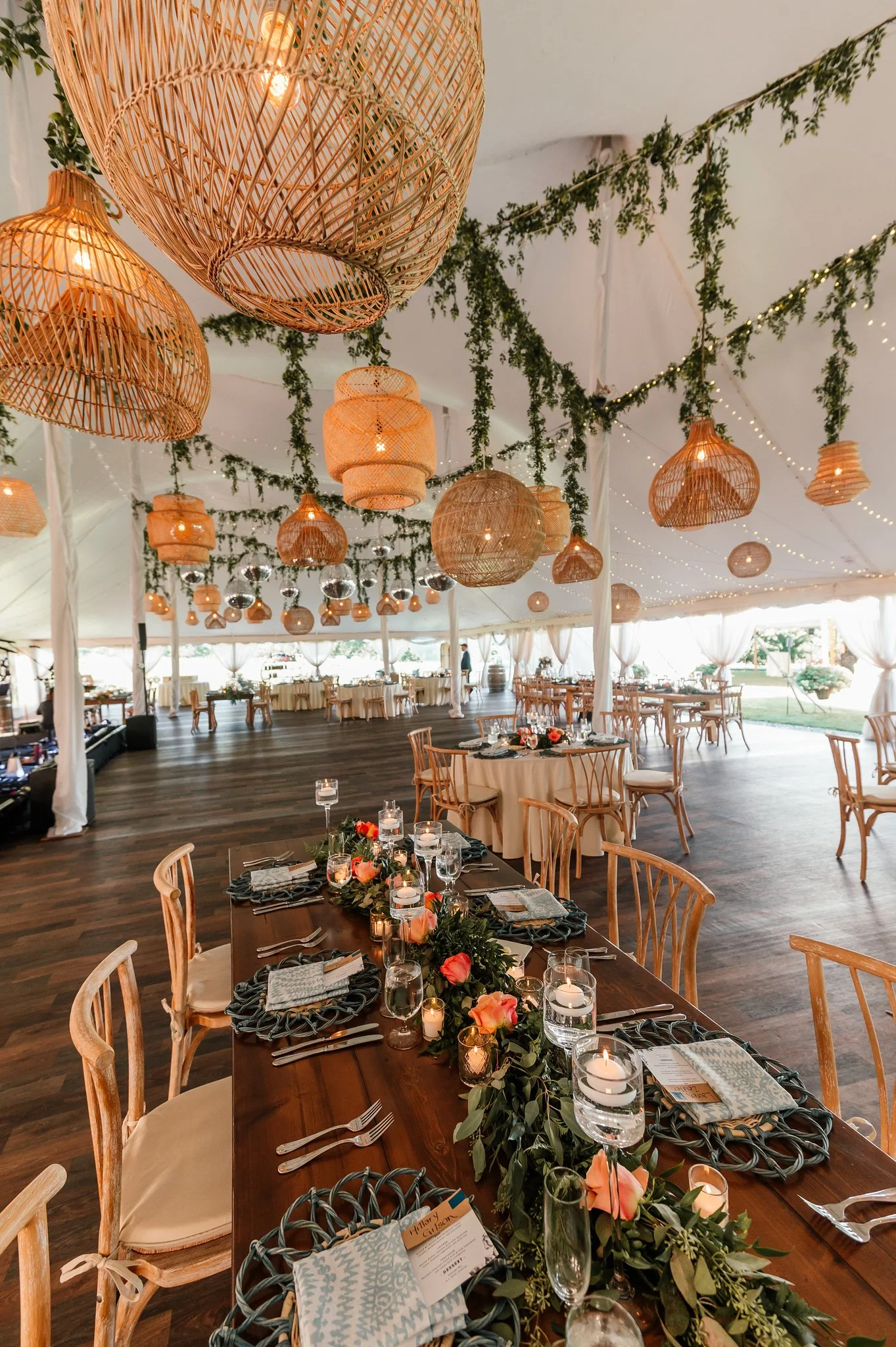 A decorated reception area with hanging wicker lamps, green garlands, and string lights, with tables set for a celebration.