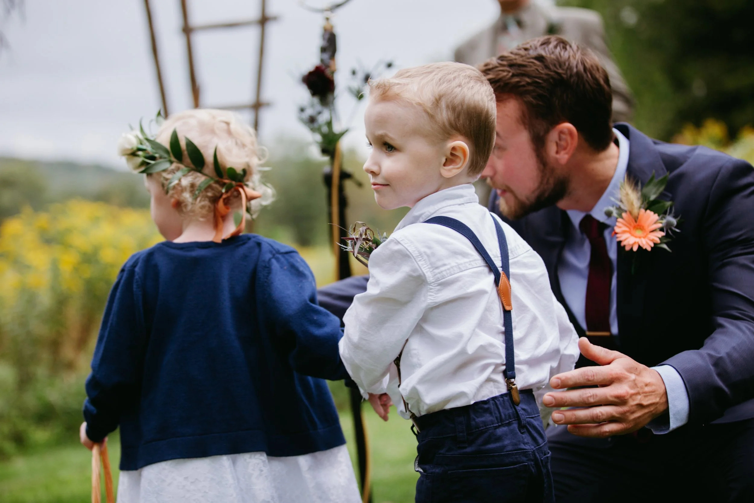 A man in a suit with a pink flower boutonniere leans down and talks to two young children, a girl and a boy, outside during daytime. The girl has curly blonde hair and wears a floral crown, while the boy has short, light brown hair and is dressed in 