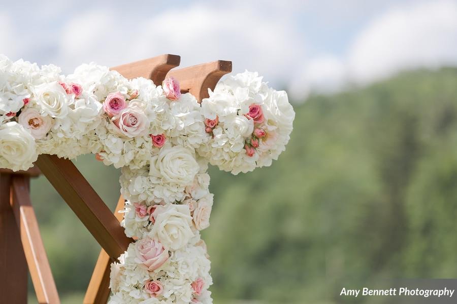 A wooden arch decorated with white and pink roses and hydrangeas, set against a blurred outdoor background, likely for a wedding ceremony.