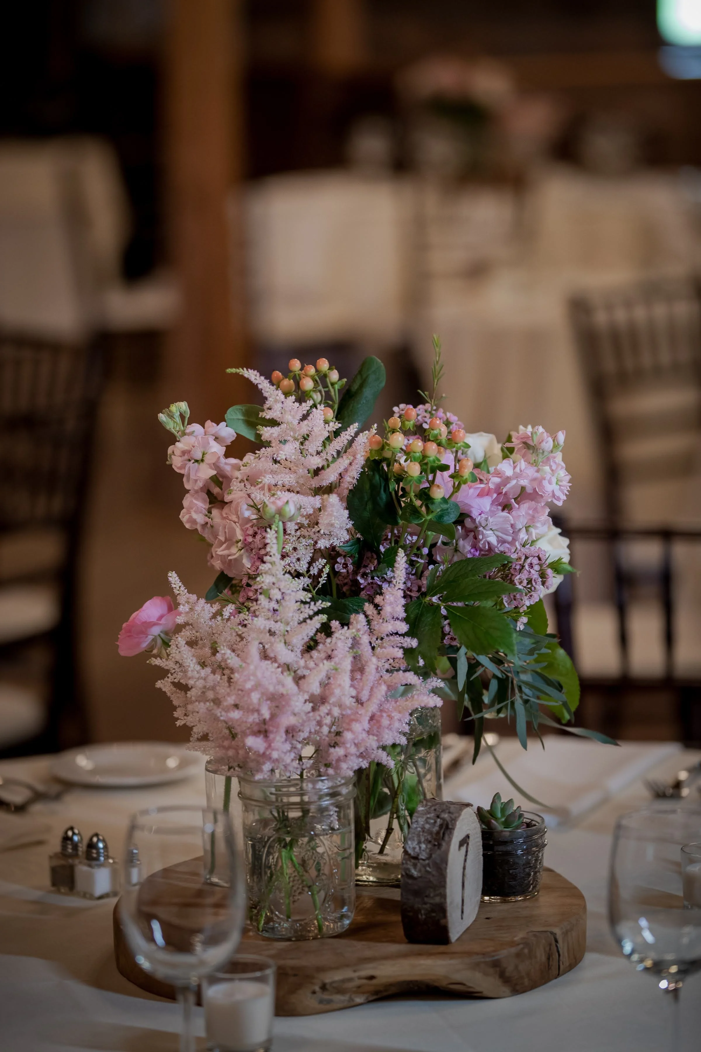 A floral centerpiece with pink and purple flowers arranged in glass jars on a rustic wooden tray, displayed on a table set for a formal event with candle holders and tableware.
