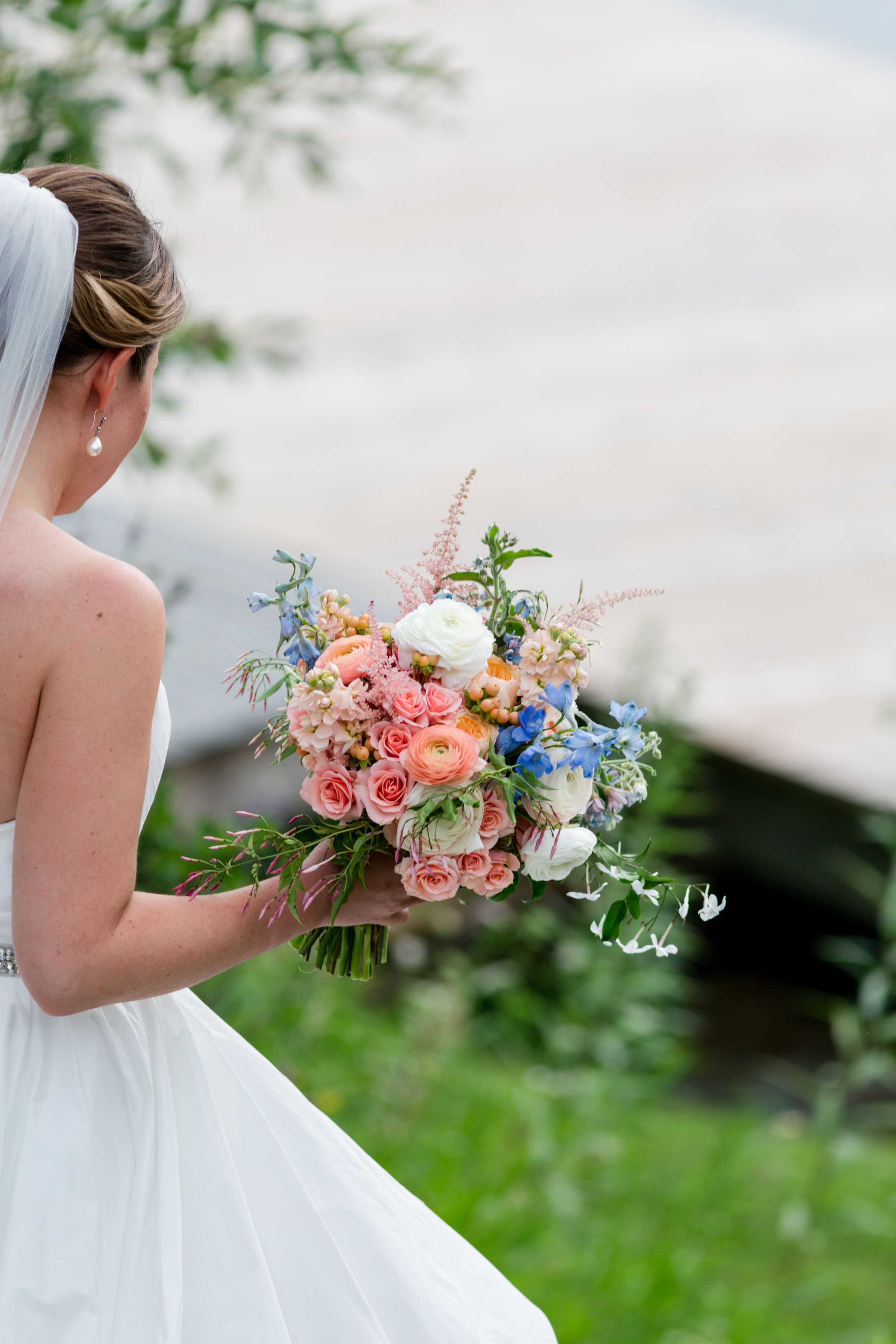 Bride with a white veil looking at a large colorful bouquet of pink, white, and blue flowers outdoors.