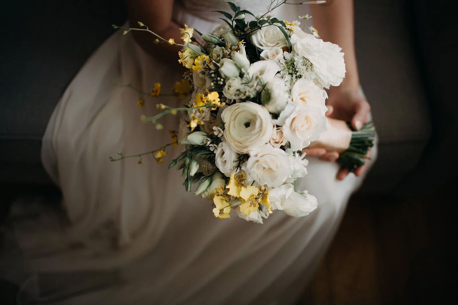A person in a beige dress holding a bouquet of white and yellow flowers, sitting on a dark sofa.