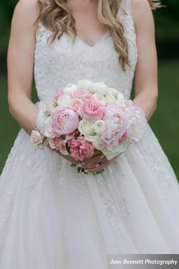 A bride in a white lace wedding dress holding a bouquet of pink and white roses outdoors.