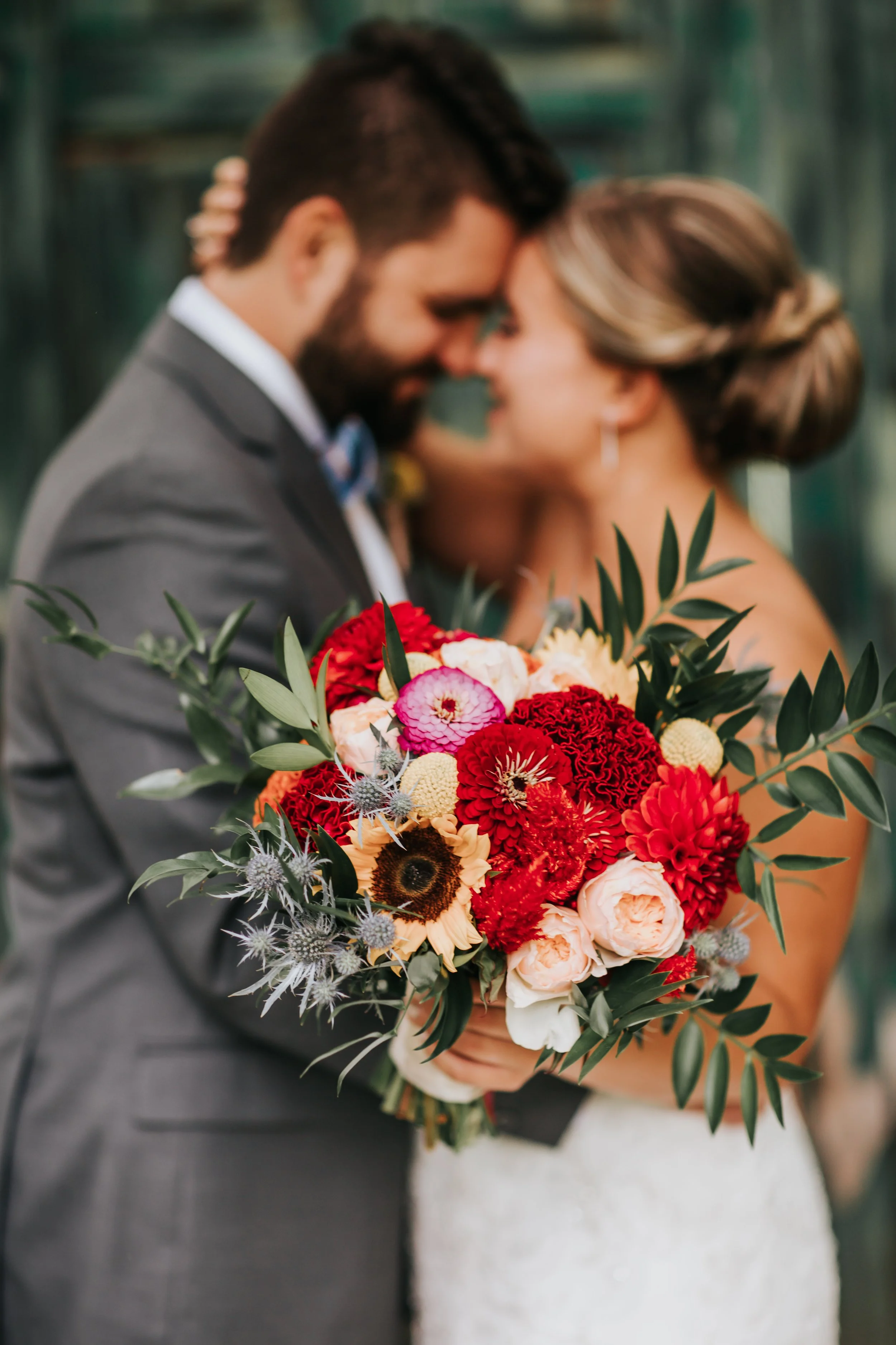 A close-up of a bride and groom touching foreheads, with the bride holding a colorful bouquet of flowers in front of them.