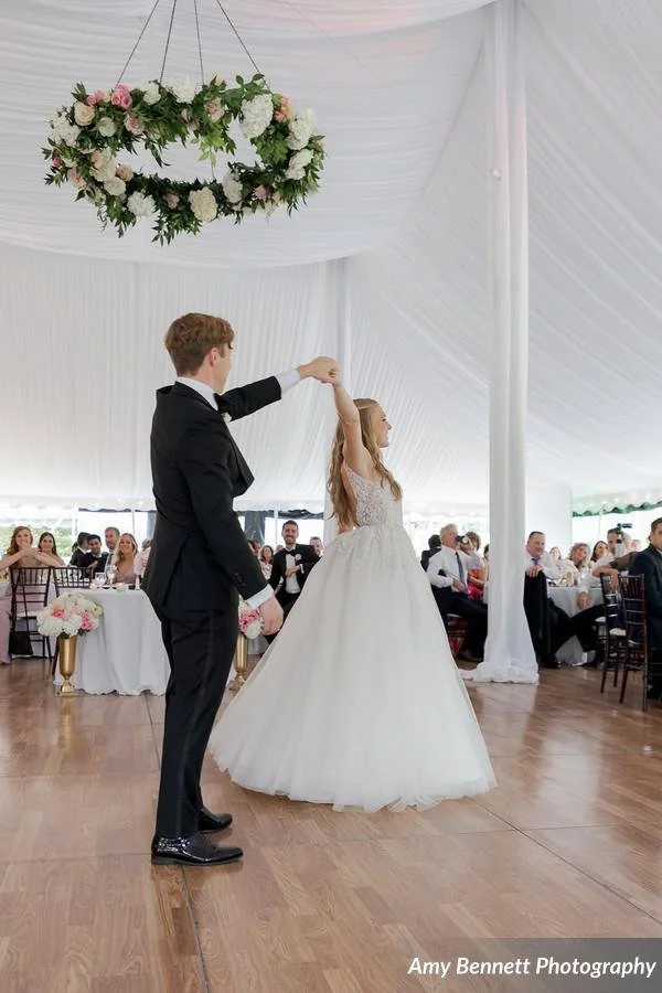 A bride and groom dancing at their wedding reception inside a decorated tent. The groom is twirling the bride on the dance floor, surrounded by seated guests.