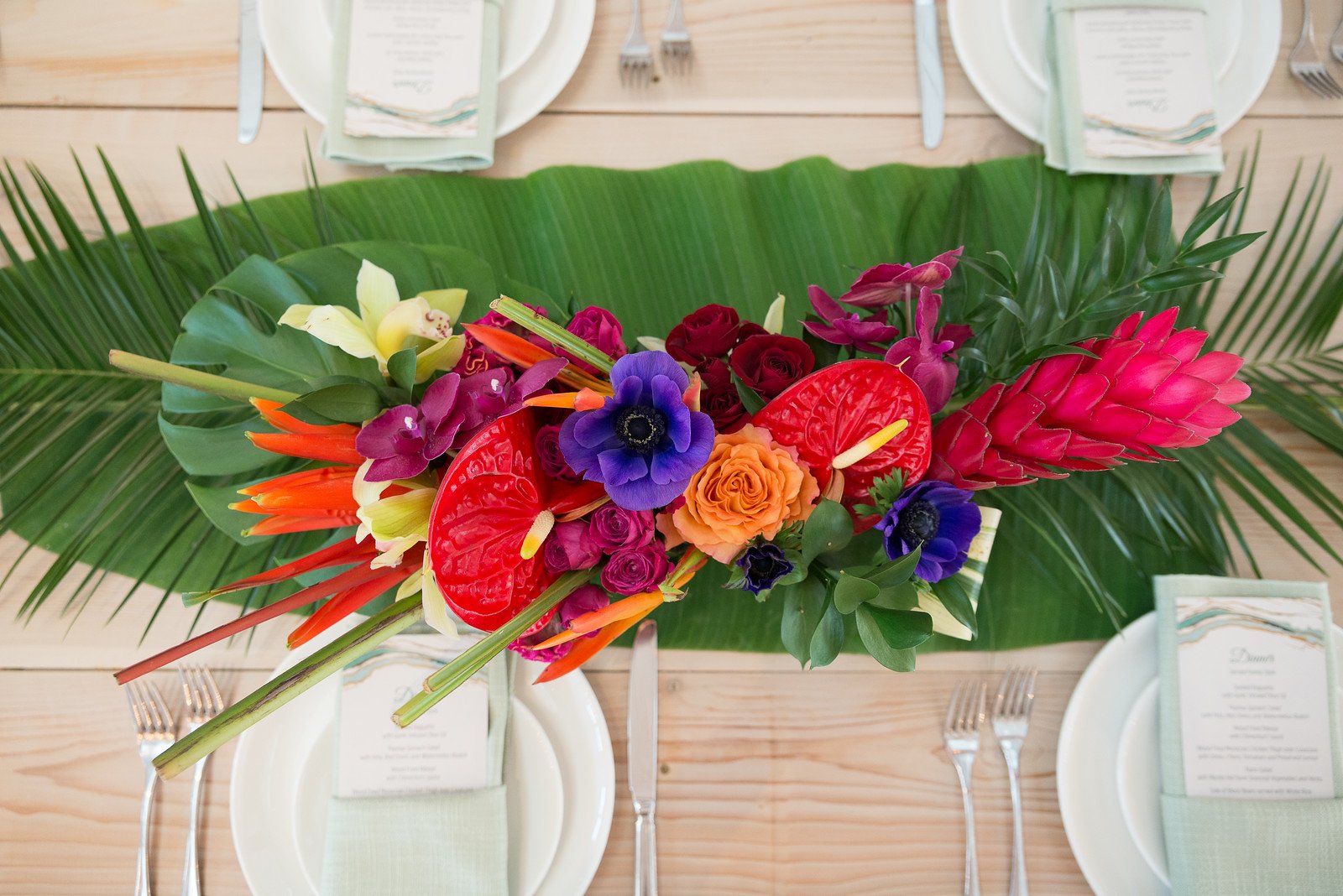 Colorful flower arrangement on a dining table with white plates, utensils, and menus, decorated with large green leaves.