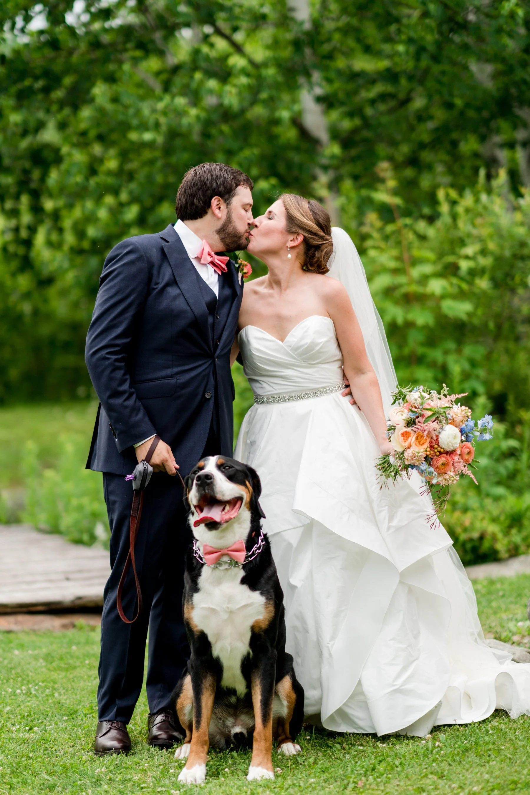 A newlywed couple kissing outdoors with a large dog in front, holding a bouquet, surrounded by green trees.