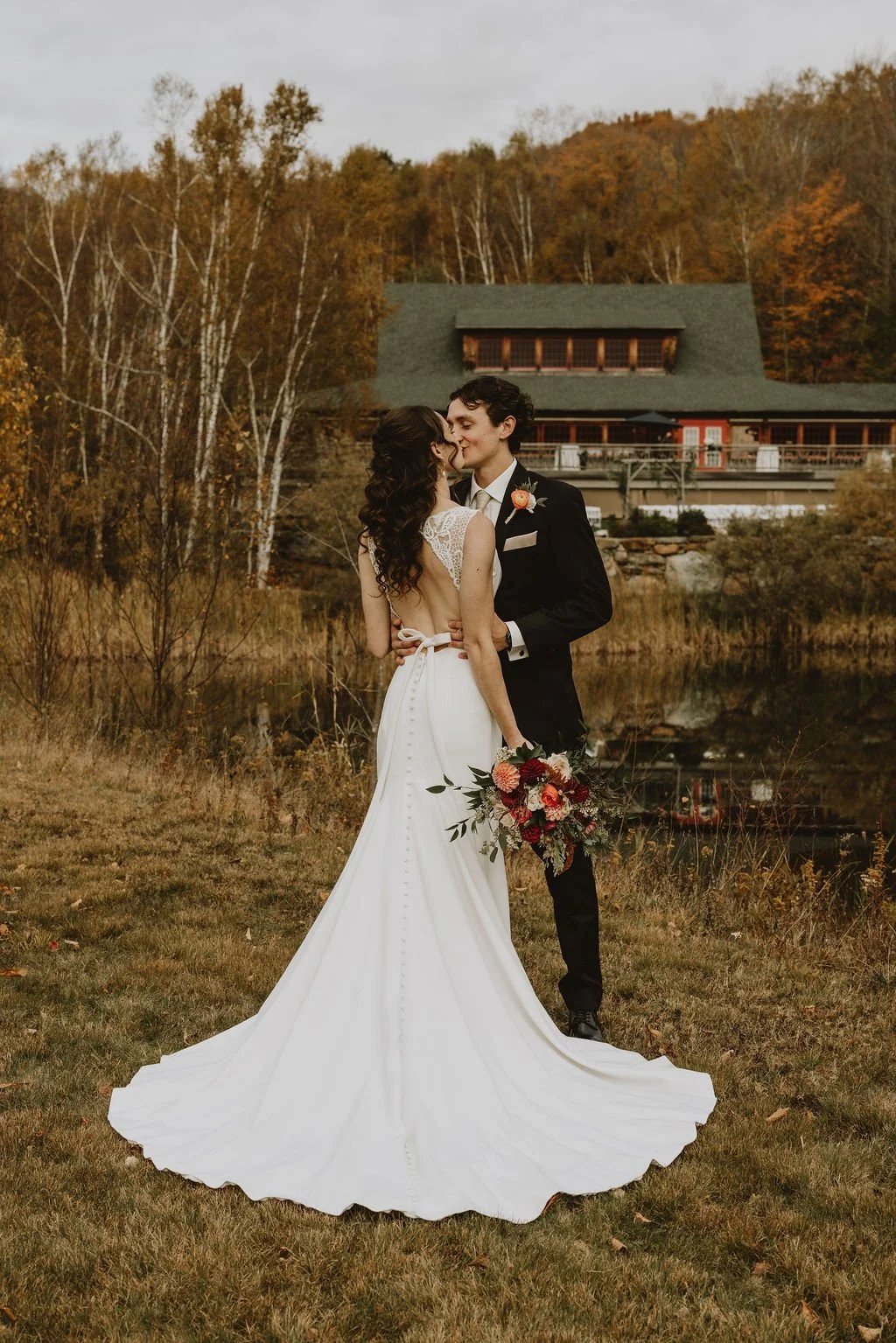 A bride and groom embrace outdoors near a pond with autumn trees and a large rustic house in the background.