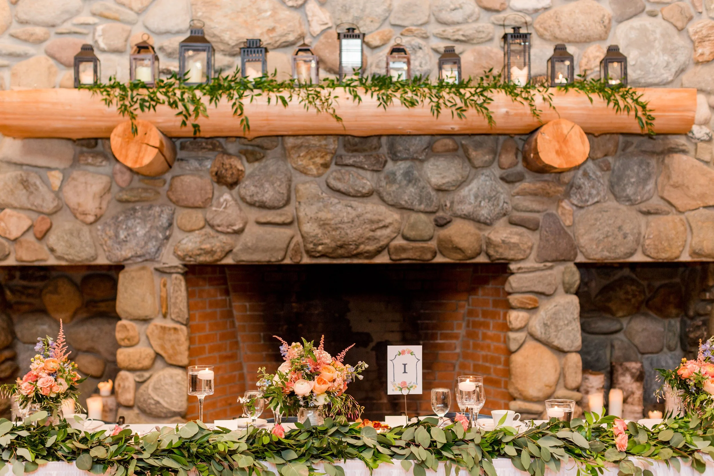 A decorated rustic fireplace mantel with candles, greenery, and lanterns, set up for a celebration or event, with floral arrangements and glasses on a table in front.