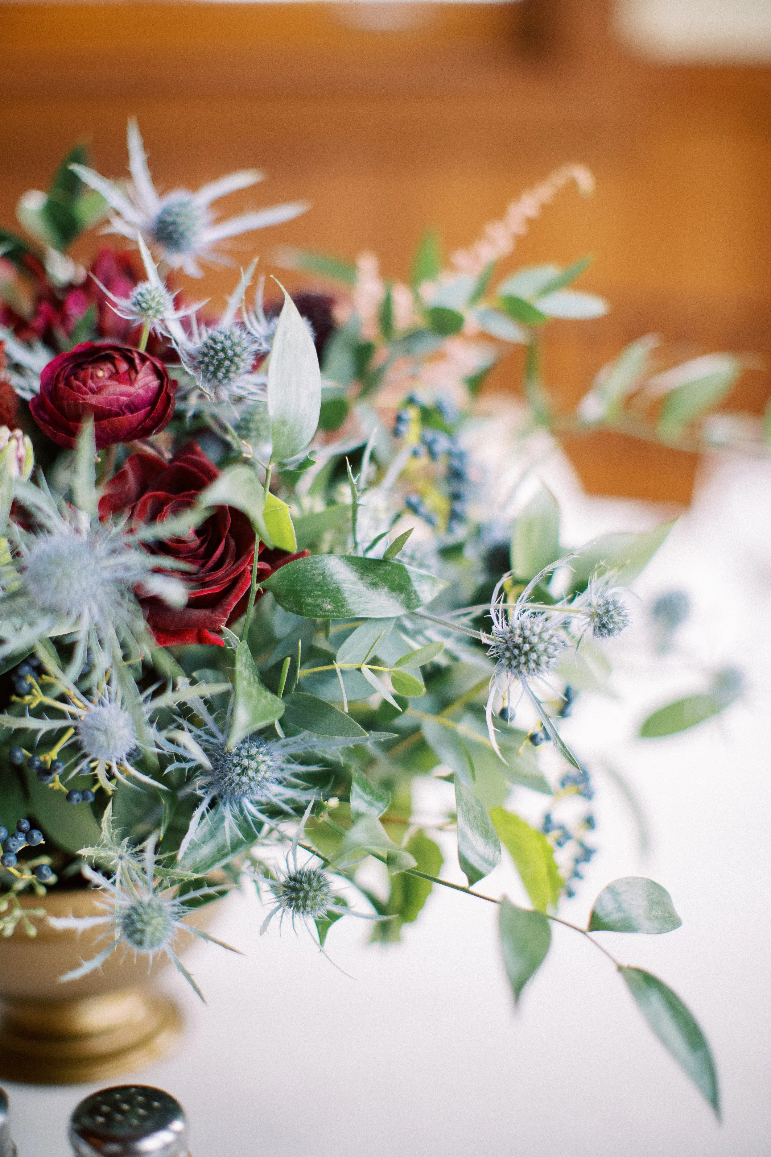 Close-up of a bouquet of flowers with various green leaves, white feathery flowers, red roses, and blue thistles on a wooden surface.