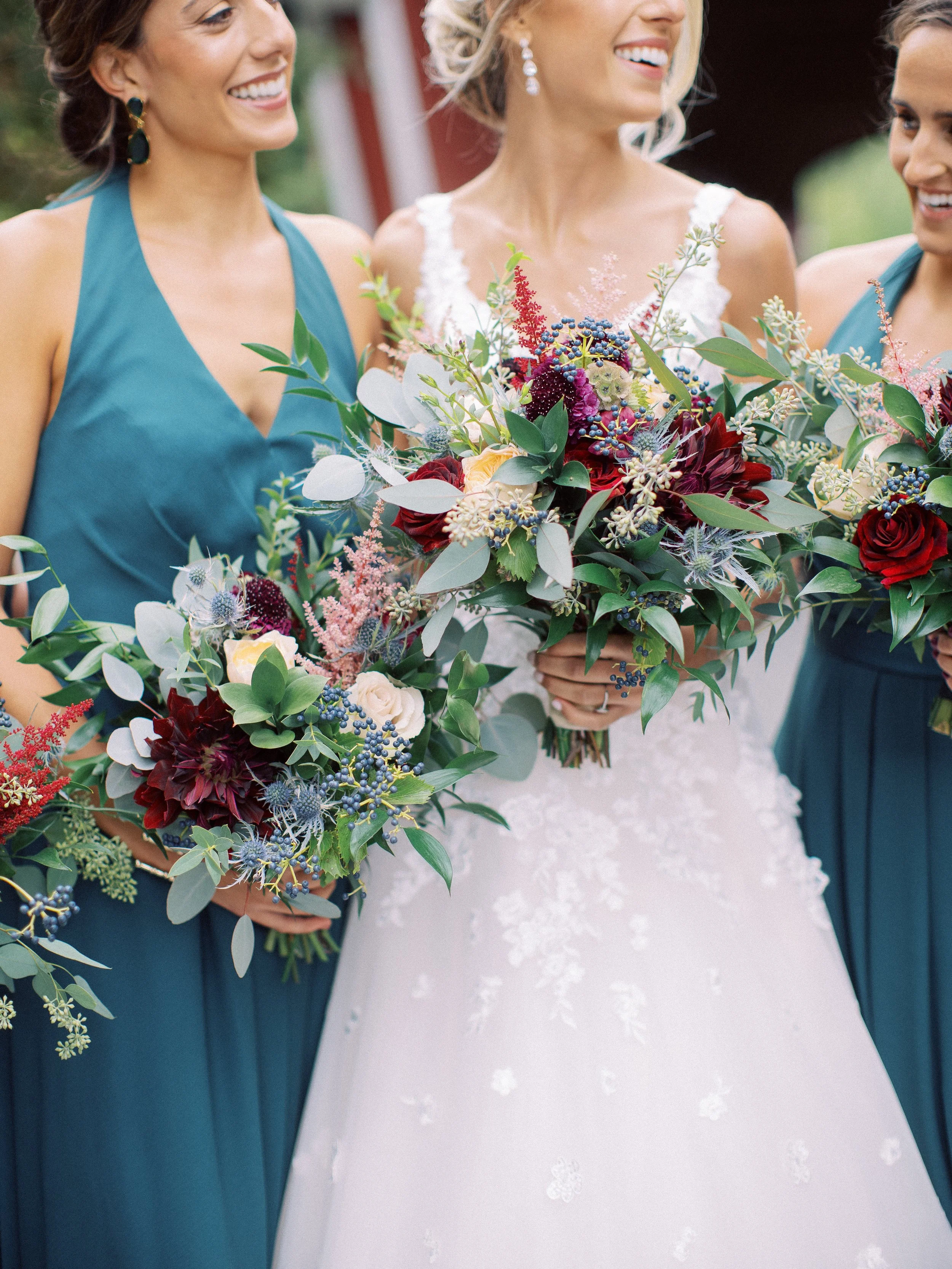 Close-up of three women at a wedding, two bridesmaids in teal dresses holding bouquets with dark red, white, pink, and purple flowers, and a bride in a white lace wedding gown.