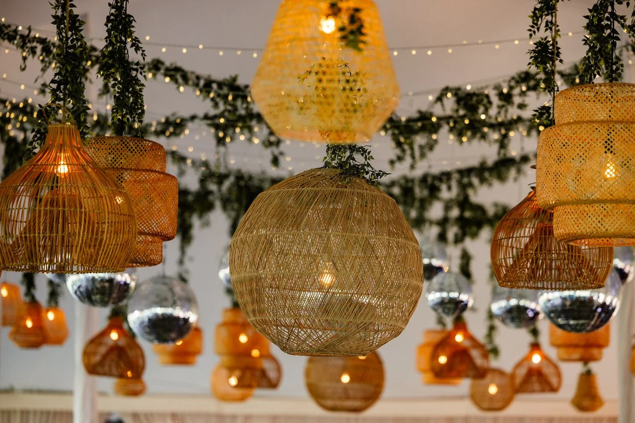 Decorative hanging wicker and bamboo lanterns and string lights with greenery, hanging from the ceiling in an outdoor or indoor setting.