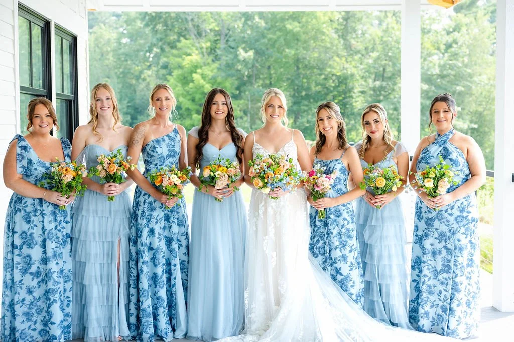 Group of women in matching blue and white dresses holding bouquets of flowers, standing indoors with large windows and greenery outside.