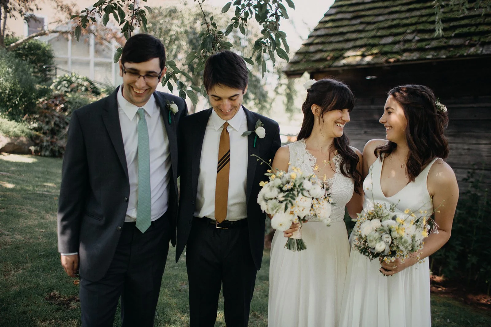 Group of five people, two men and three women, smiling and enjoying each other's company outdoors at a wedding or celebration. The women are holding bouquets of flowers. The men are dressed in suits with boutonnieres, and the women are in white dress