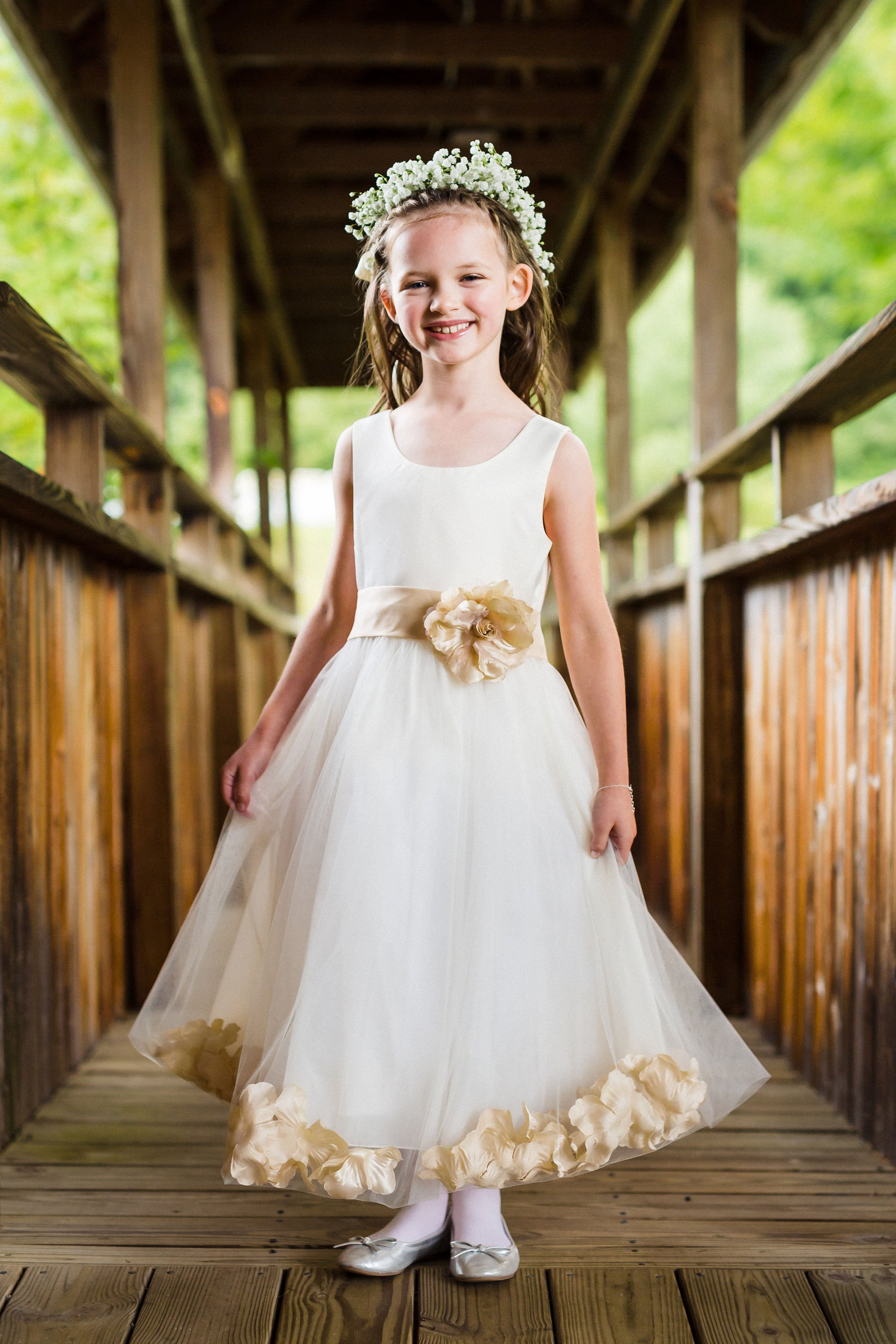 A young girl in a white dress with a floral sash, wearing a flower crown, standing on a wooden bridge with a green outdoor background, smiling.